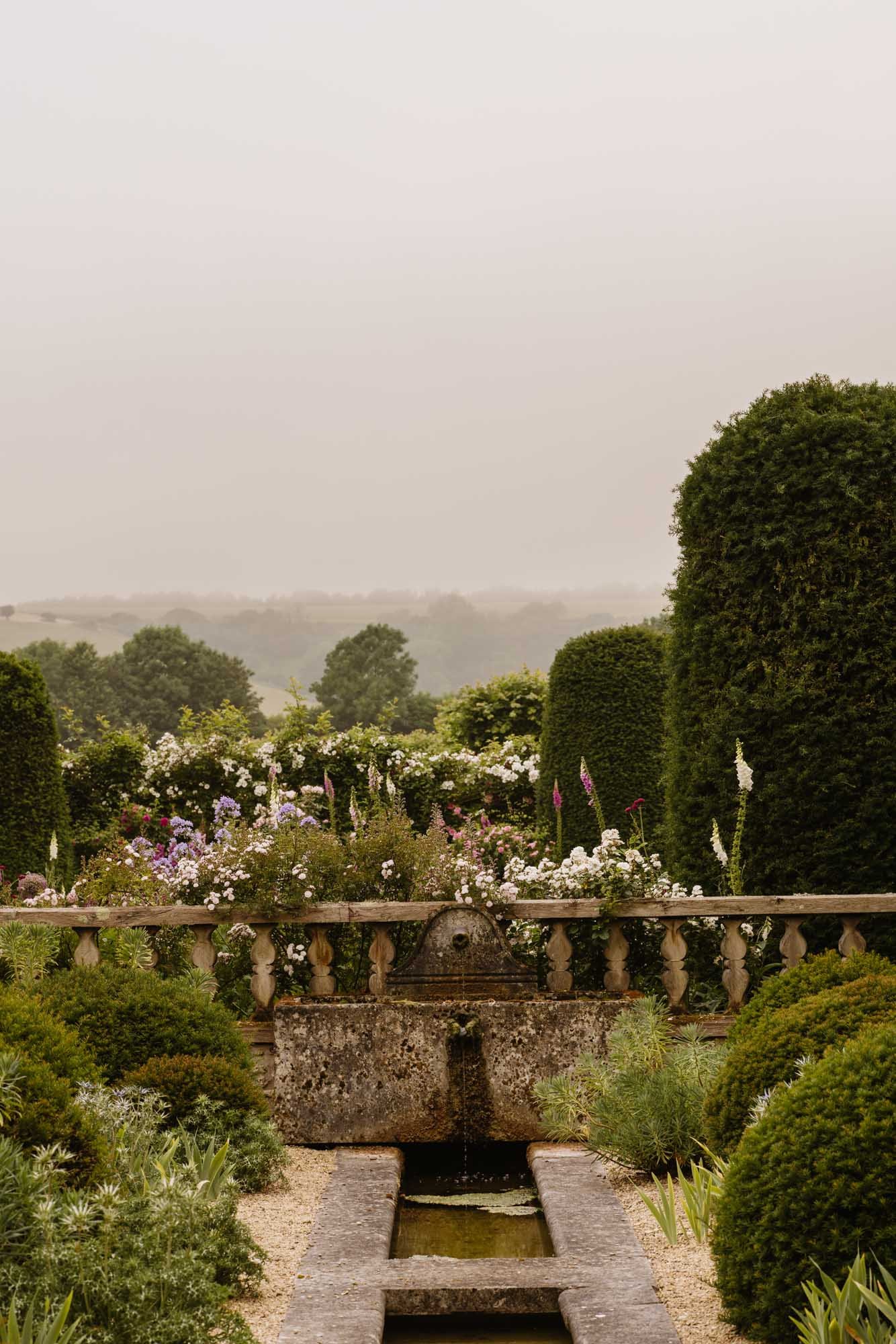 Dorset Walled Garden with a stone fountain, lush bushes, and blooming flowers, surrounded by tall green hedges under a misty sky.