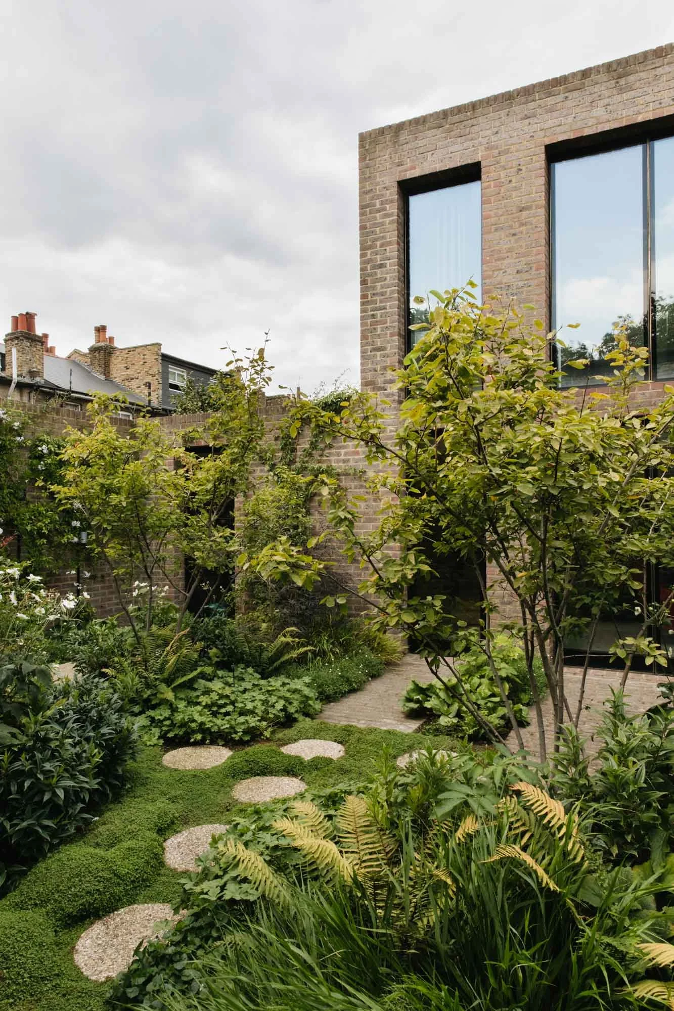 A lush garden with various green plants, trees, and a stone pathway in front of a modern brick house with large windows. Non Morris garden design
