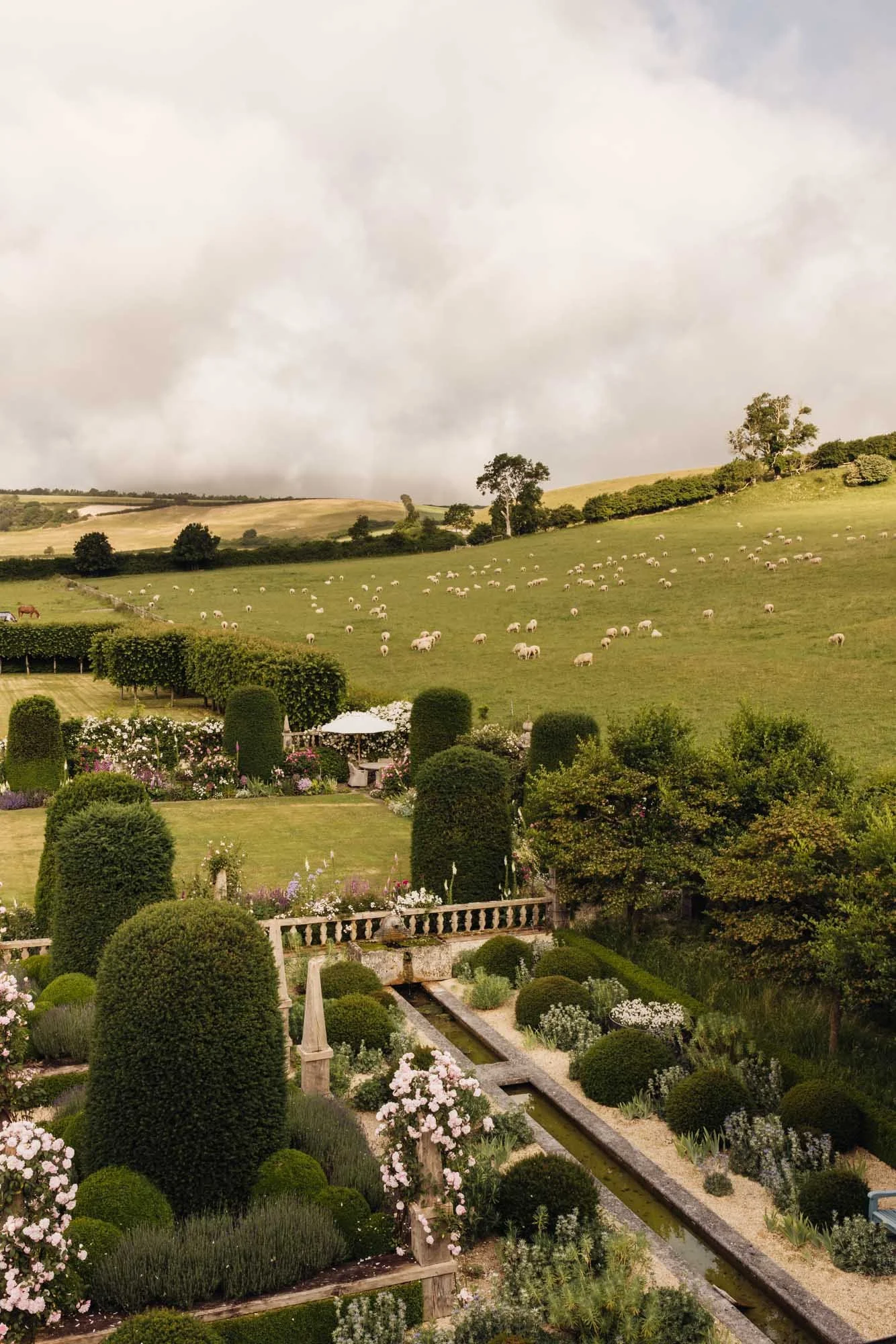 Landscape with a beautifully manicured garden in the foreground, a large grassy hill with sheep grazing, and a cloudy sky in the background. Dorset Walled Garden in Somerset