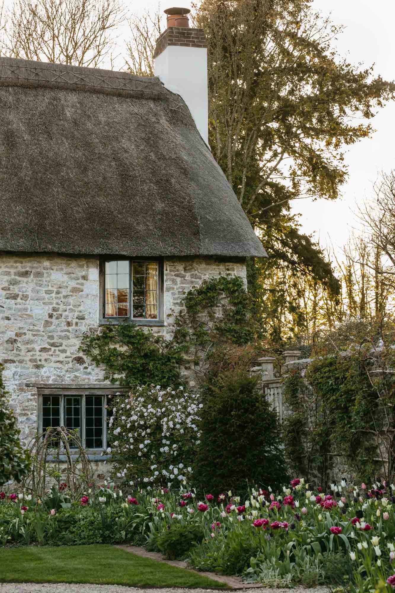 A stone cottage at South Wood Farm with a thatched roof, surrounded by a lush garden with colorful tulips and flowering bushes, and tall trees in the background.