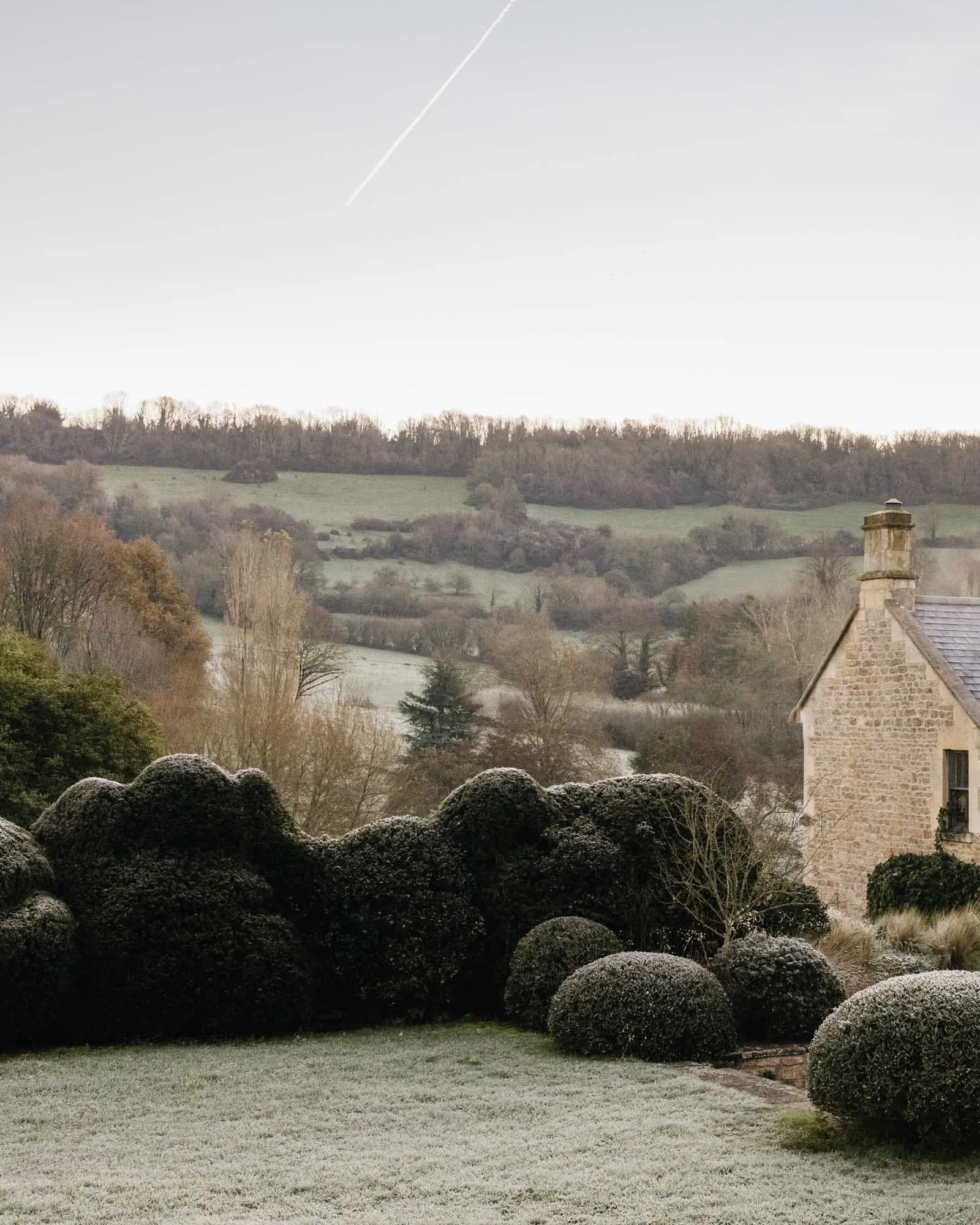 A frosty garden with round bushes and a stone house in the background, overlooking rolling hills and trees, under a clear sky with a jet contrail.