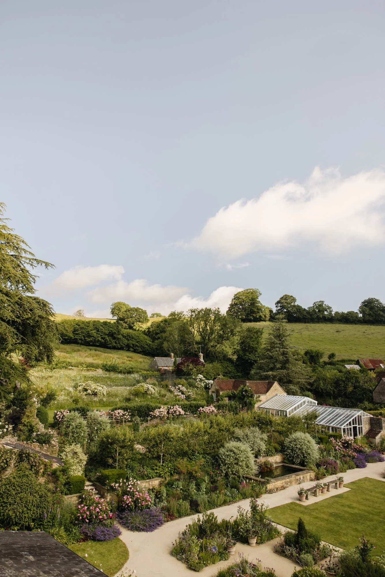 Batcome House and Gardens with a scenic view of a lush garden with colorful flowers, a lawn, and a greenhouse, set against hilly green landscape and a blue sky with scattered clouds.
