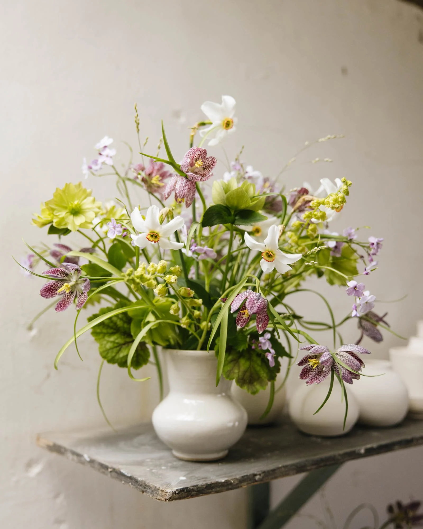 Green and Gorgeous white vase filled with fresh narcissi and fritillaria, including white, pink, and purple blooms, sitting on a rustic wooden shelf.