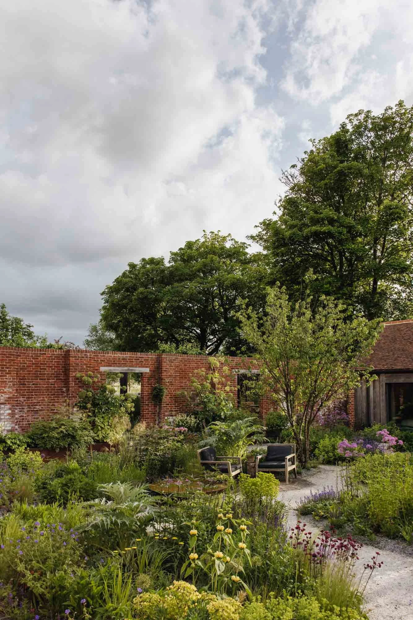 Oxfordshire Country Garden with various plants and flowers, a brick wall with a small opening, and outdoor seating under trees, with a partly cloudy sky overhead.