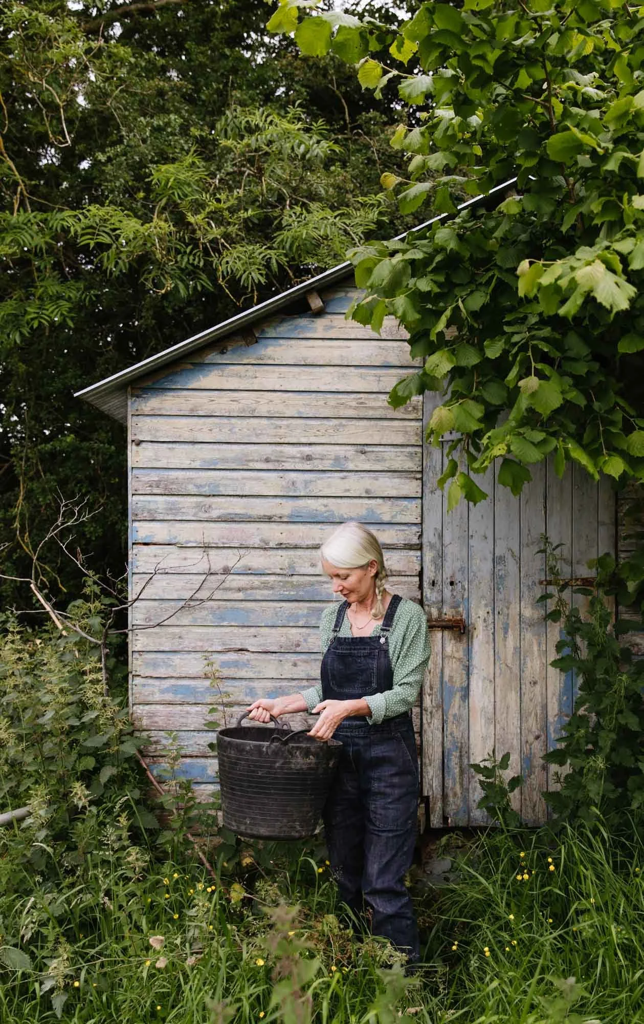 Alison Jenkins with long gray hair, wearing dark overalls and a green polka dot shirt, stands outdoors holding a black bucket near a weathered wooden shed surrounded by green plants and overgrown grass.