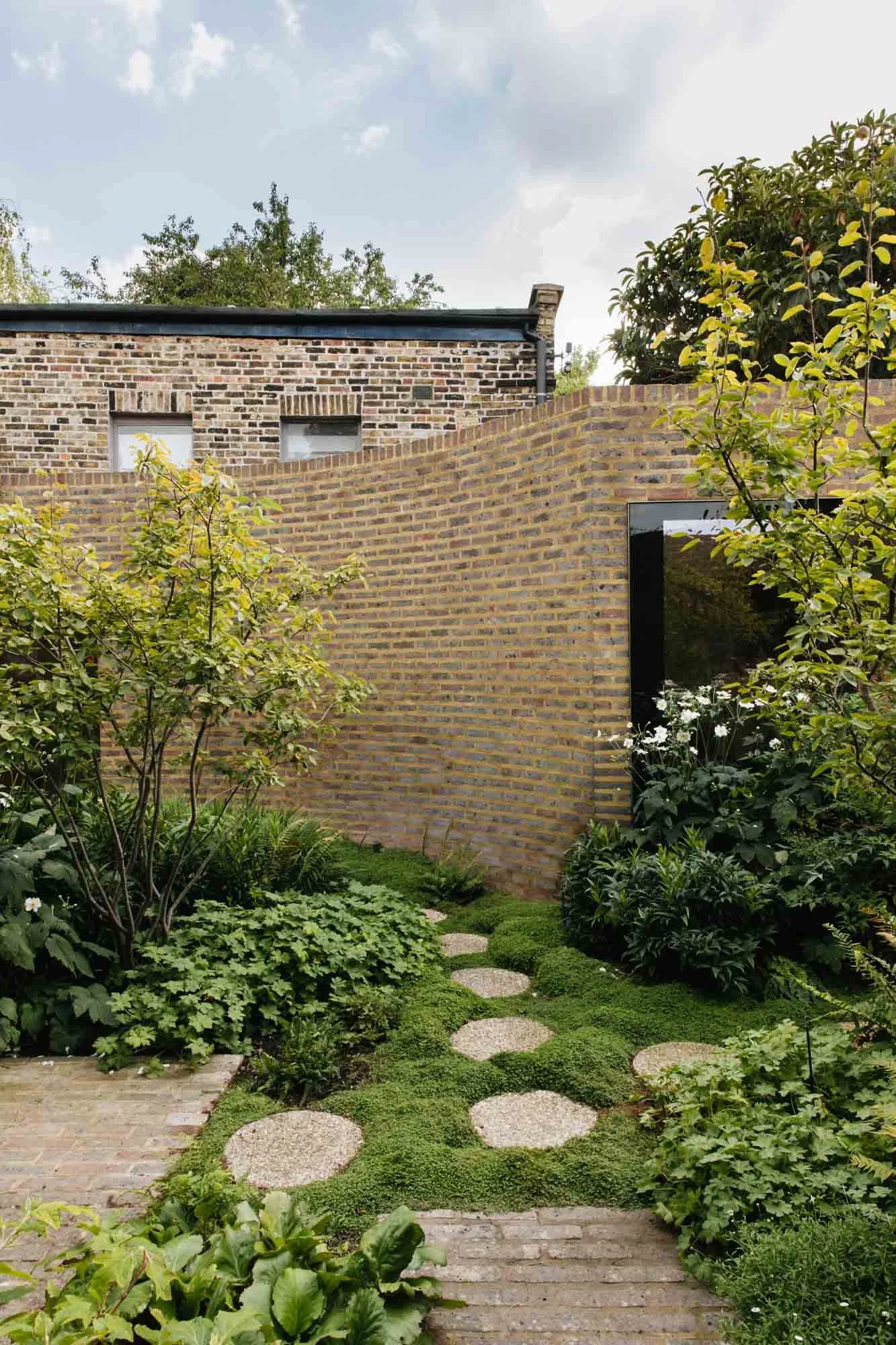 A lush garden pathway with stepping stones, surrounded by green plants and bushes, with a brick wall on the right and a taller brick building in the background under a partly cloudy sky. Non Morris garden design