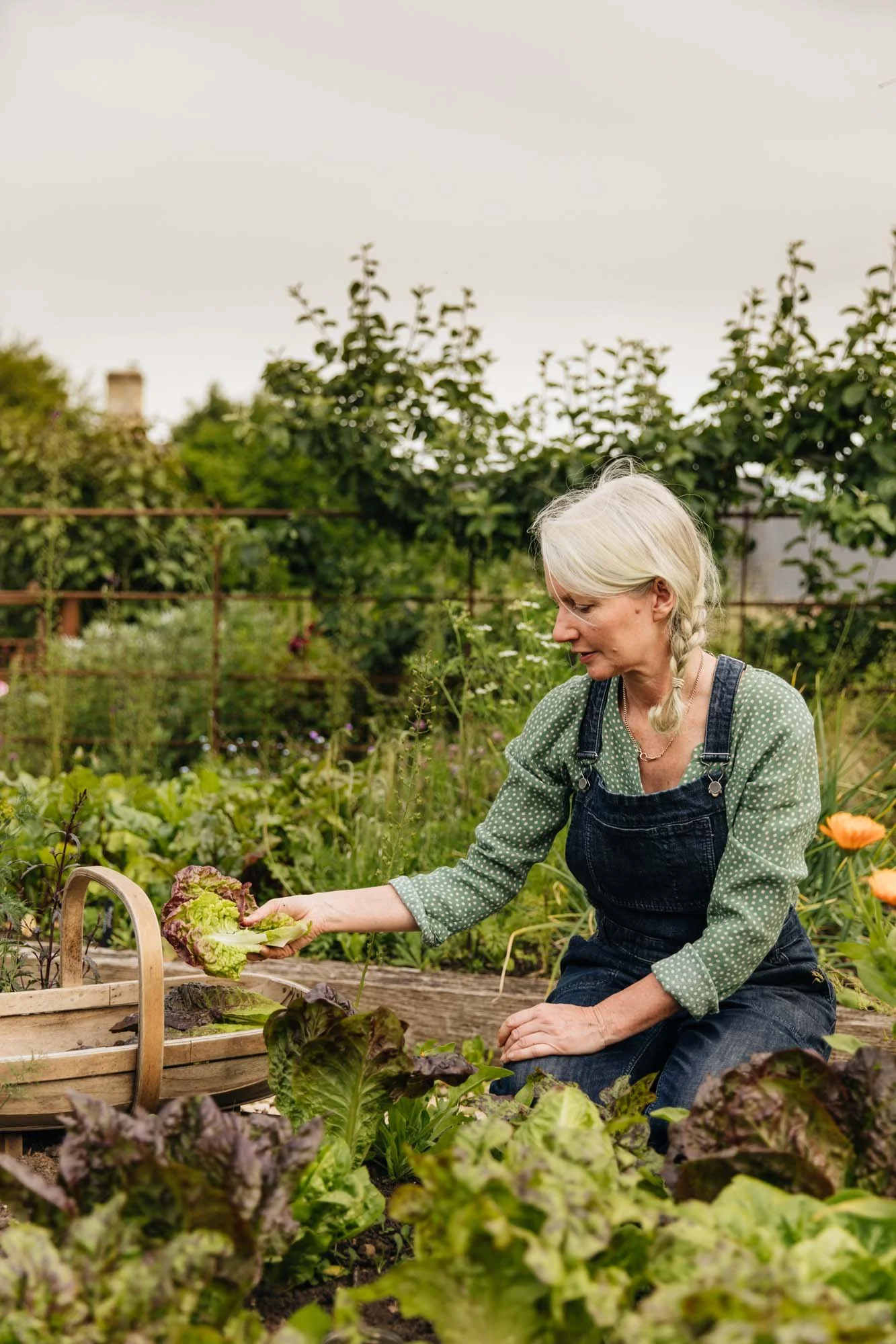 Alison Jenkins in a braid wearing a green polka-dot shirt and denim overalls gardening in a lush vegetable garden Damson Farm