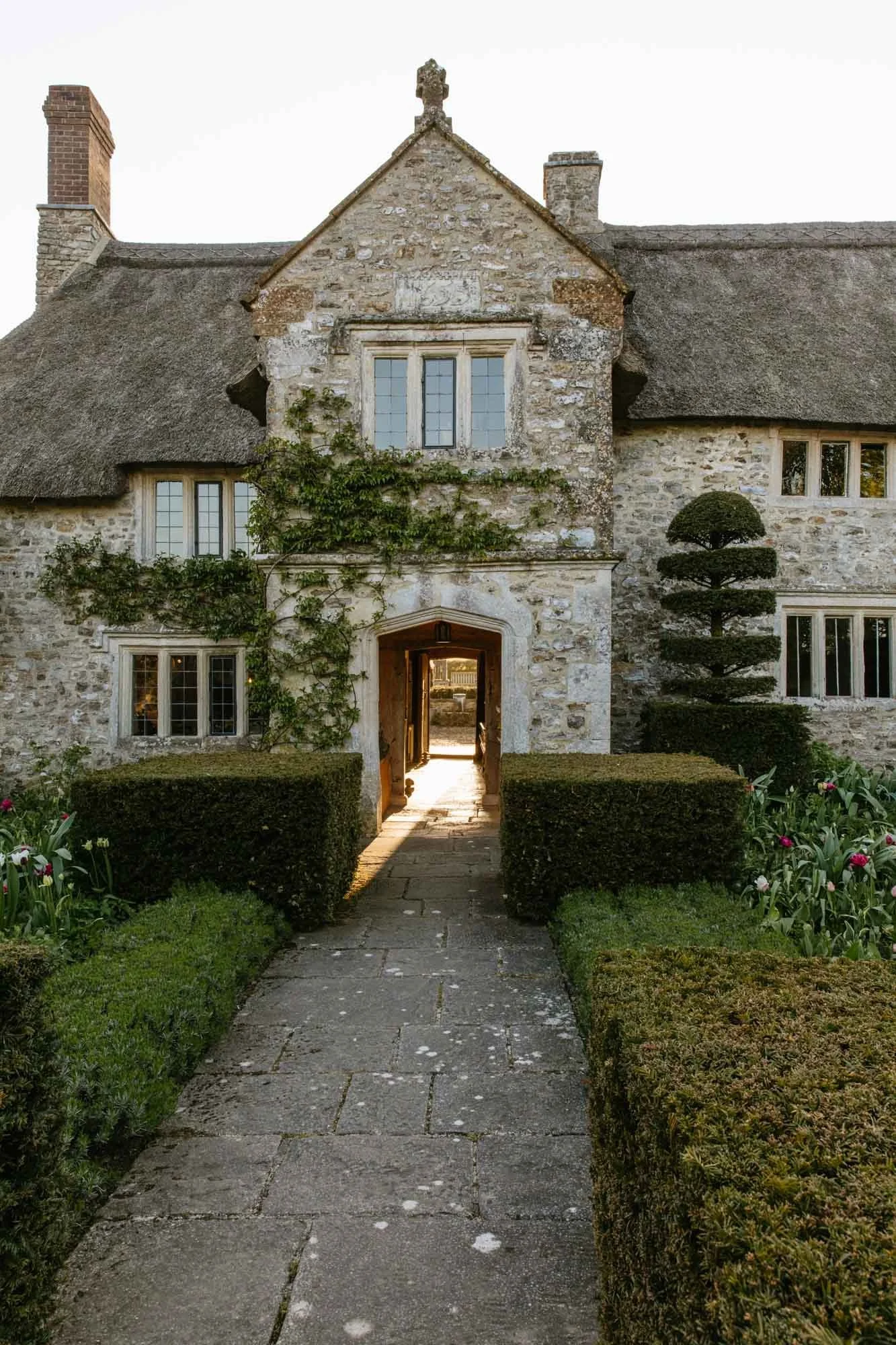 Front view of South Wood Farm with a thatched roof, ivy growing on the wall, and a neat garden with trimmed bushes and colorful flowers.