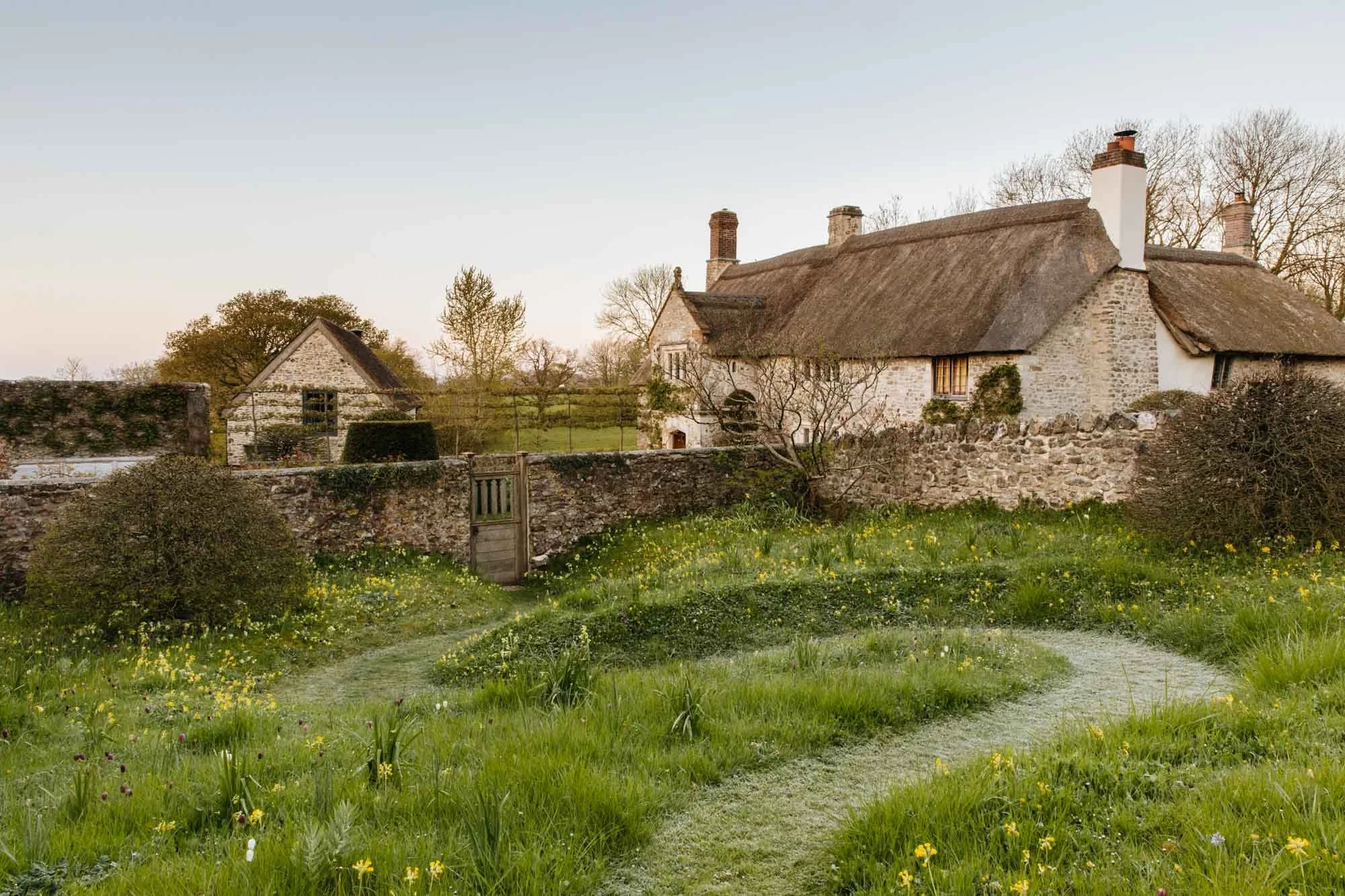 South Wood Farm garden with view of the cottage with a thatched roof, surrounded by a stone wall, green grass, and small bushes, under a clear sky.