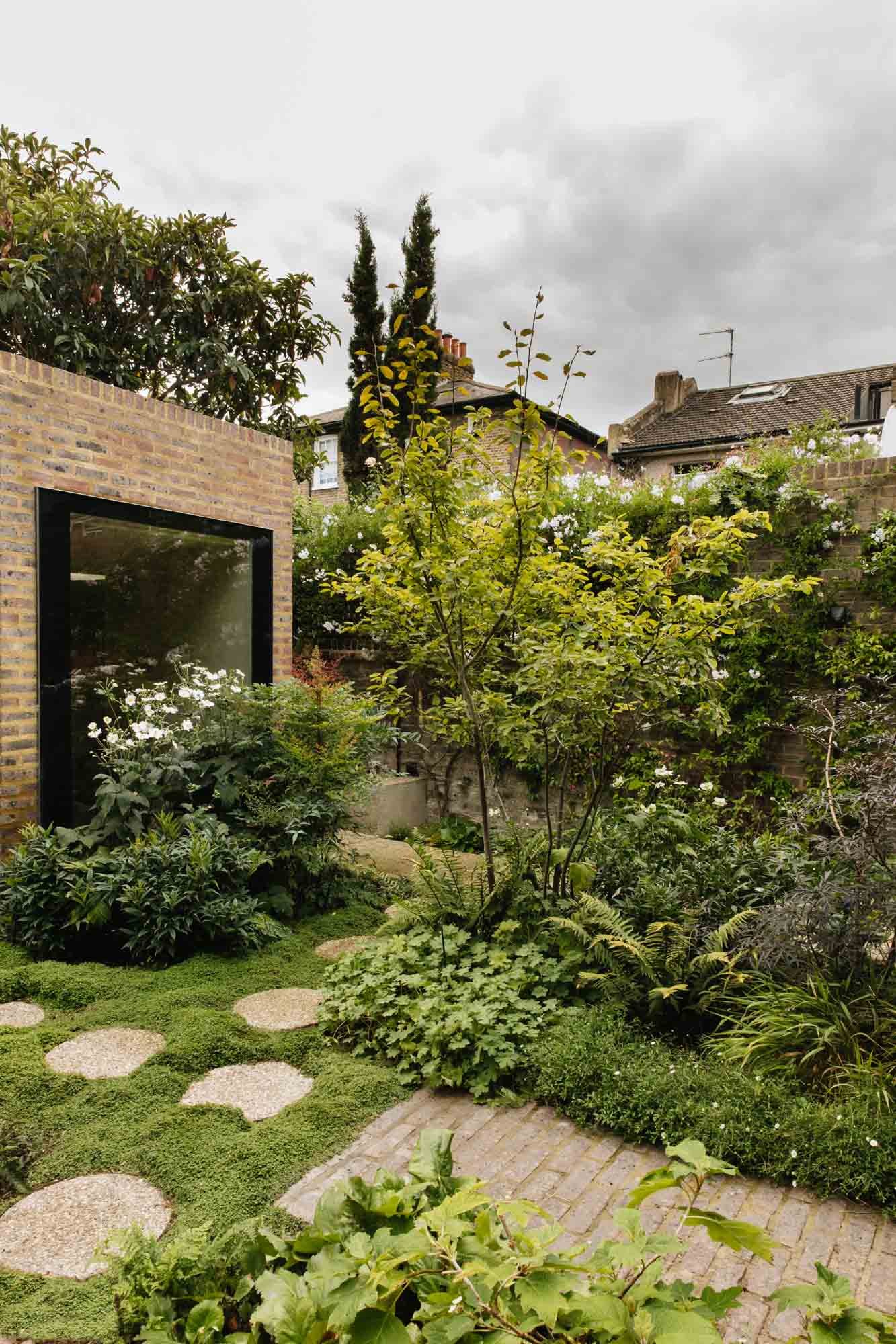 A lush garden with a stepping stone path, dense green foliage, flowering plants, and a brick wall with a large mirror or window reflecting plants, under a cloudy sky. Non Morris garden design