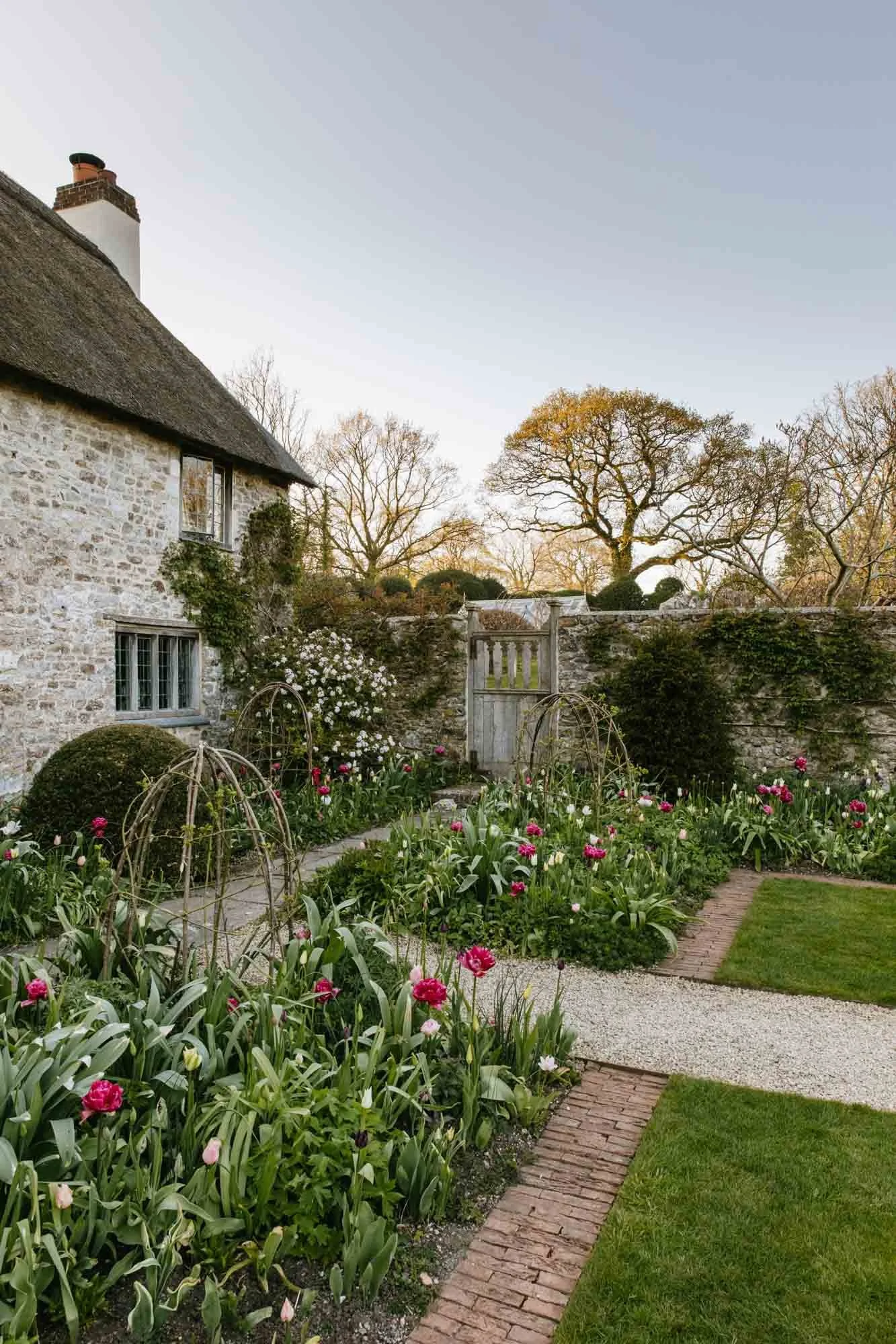 South Wood Farm garden with tulips, a stone house with small windows, a brick pathway, and trees in the background during late afternoon or early evening.