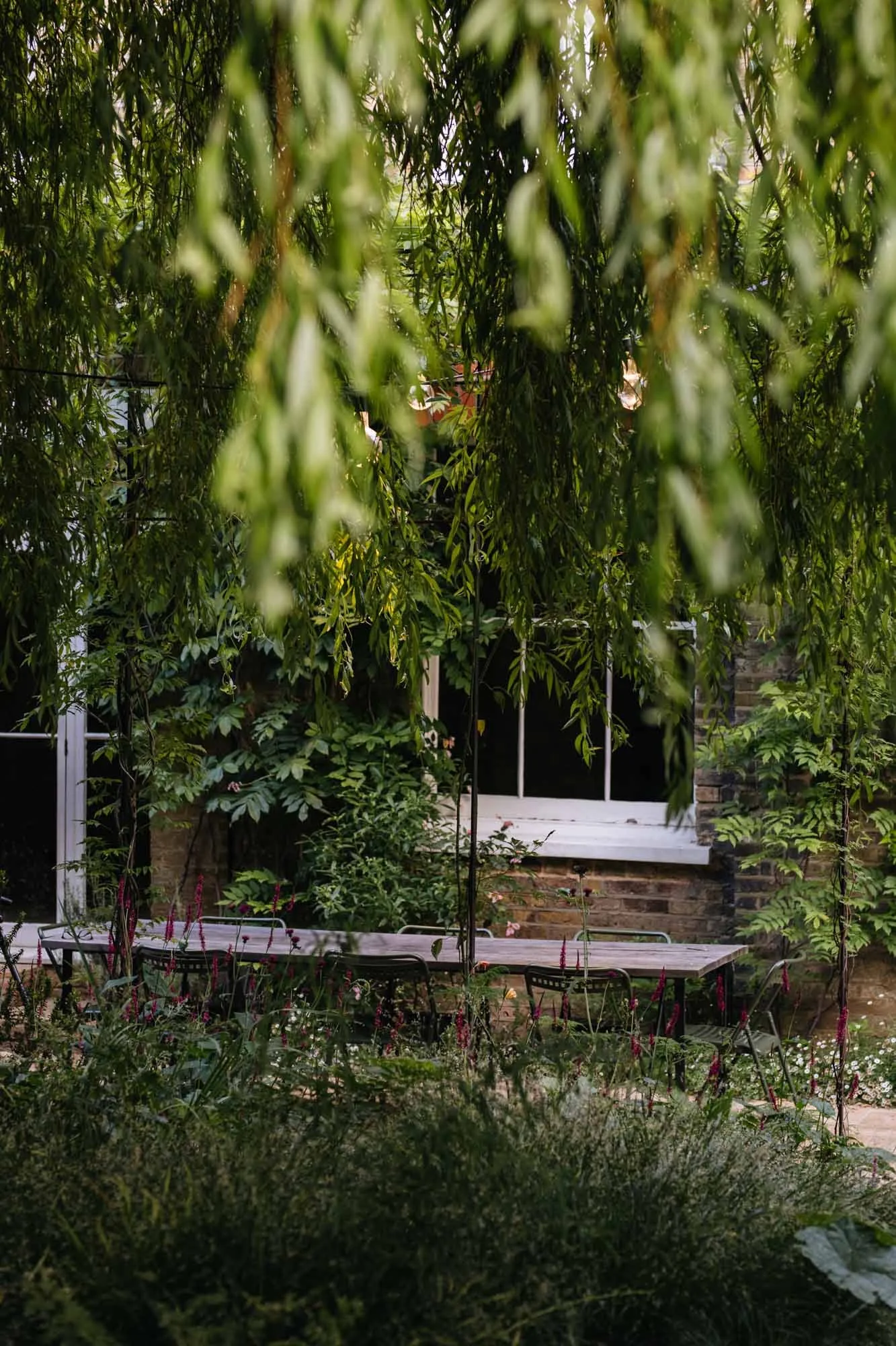 A garden scene with hanging vines and leaves obscuring a view of a house's window and an outdoor table with chairs. Sarah Price designed garden