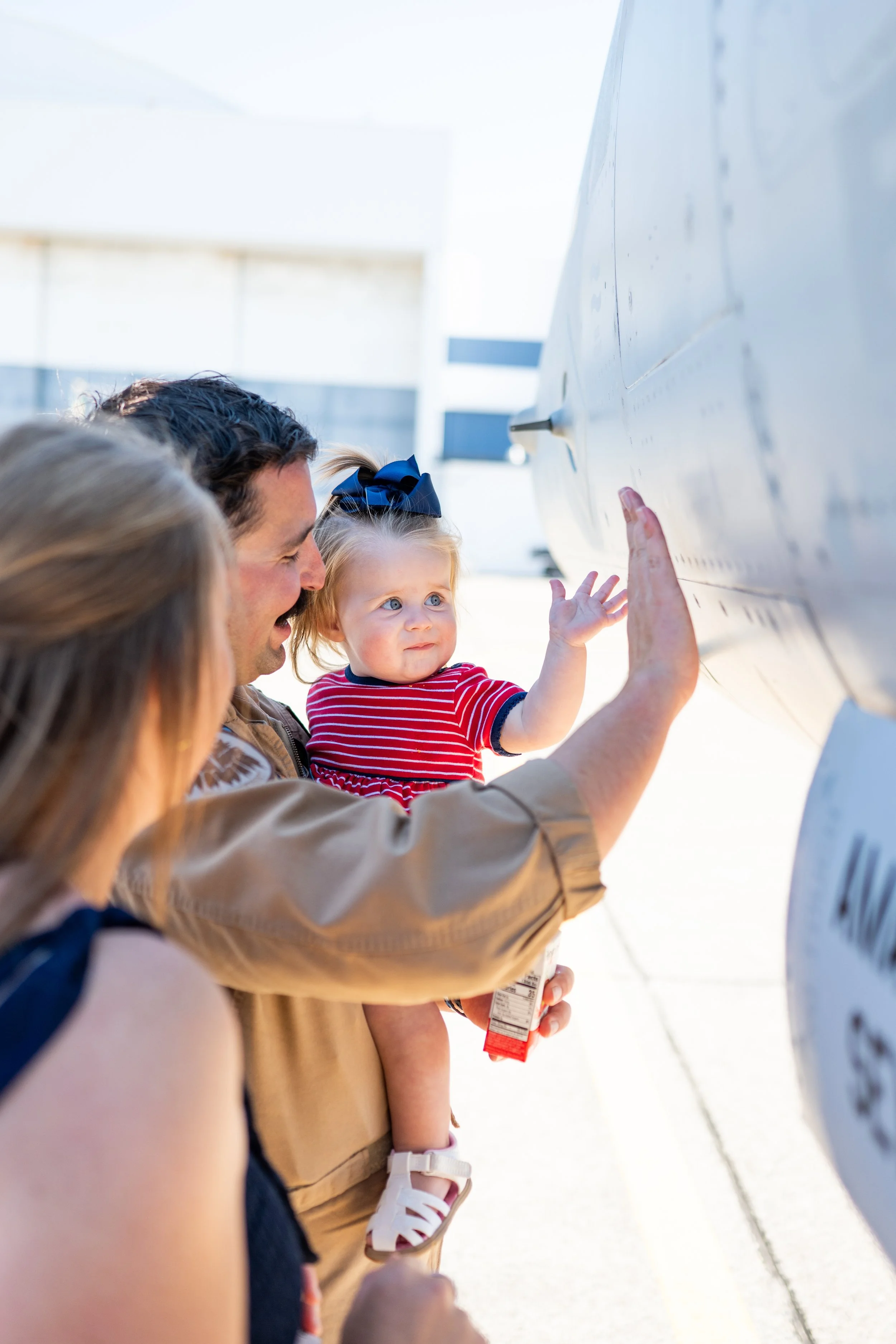 A man holding a young girl with blonde hair and a blue bow, pointing at a white airplane, with the presence of a woman partially visible in the foreground.
