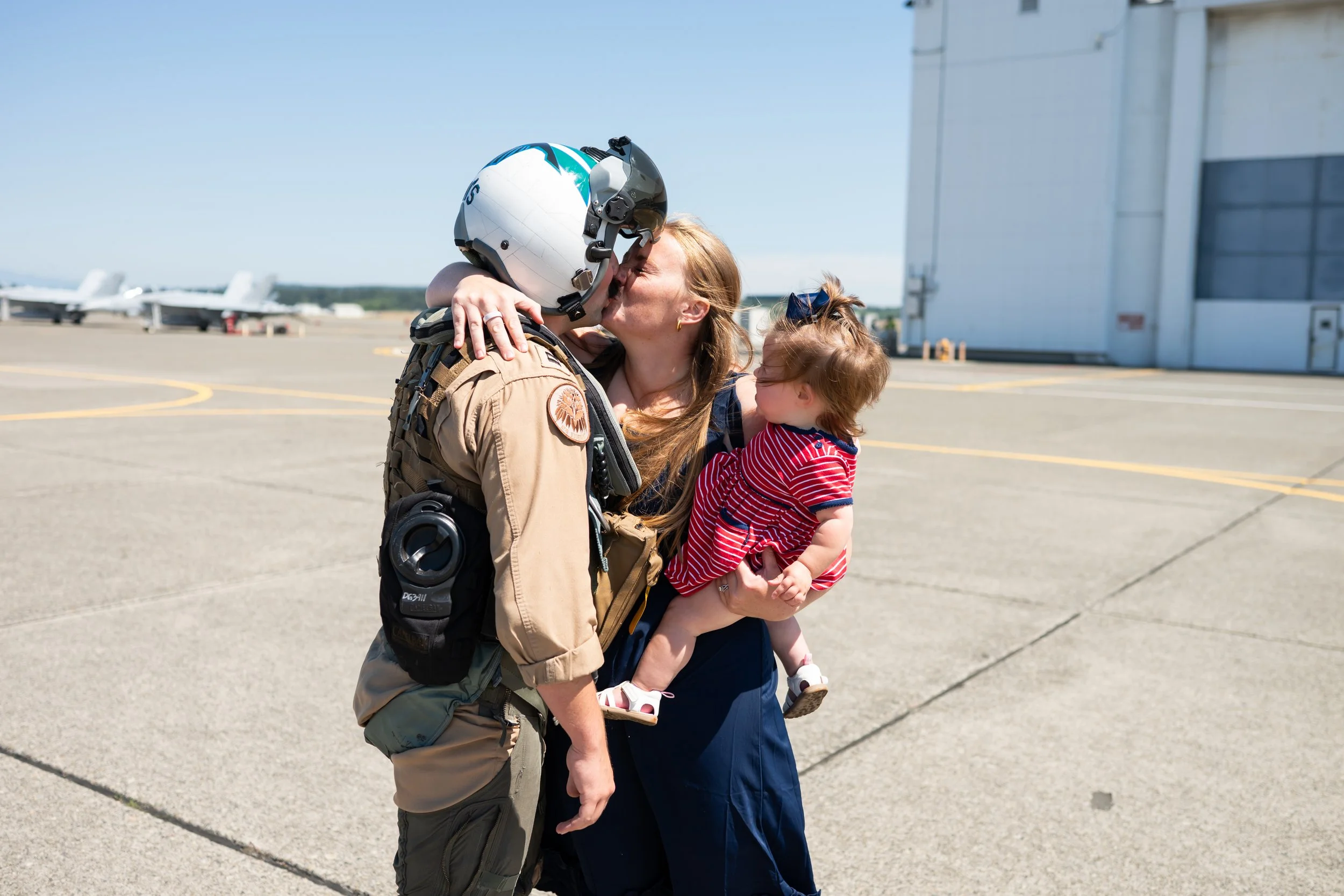 A soldier in uniform embracing a woman and a young girl on an airport tarmac, with private jets in the background.