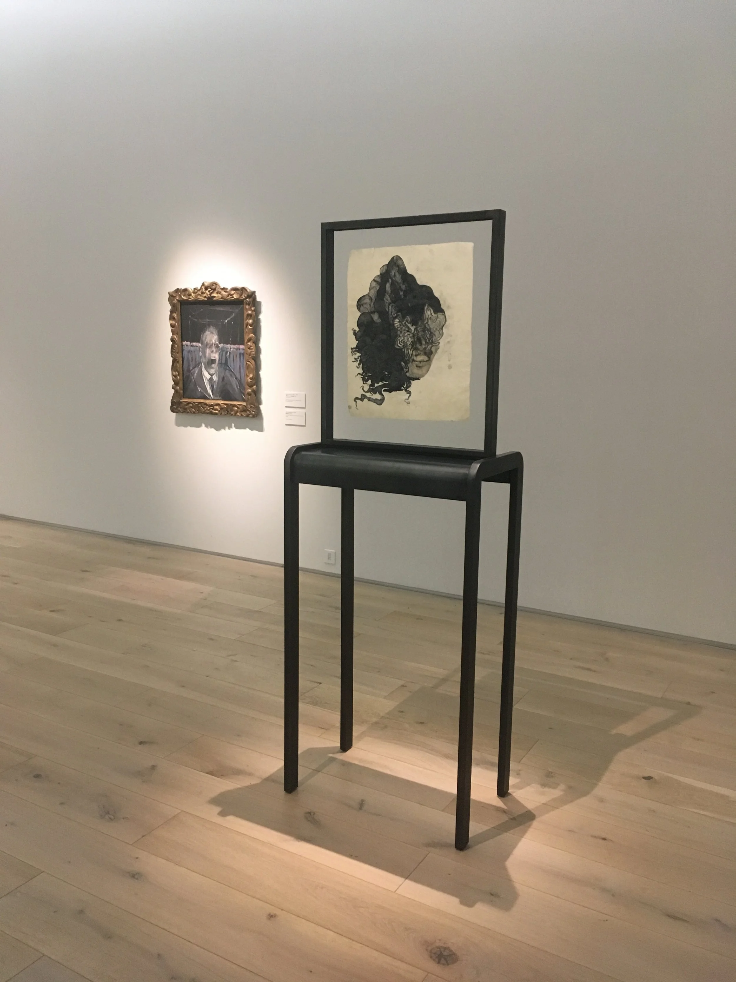  Image description: A view of two artworks in a white cube gallery space: in the foreground, a semi-figurative work on paper resembling a face in the process of transformation, pressed between glass in a freestanding clean-lined black metal structure