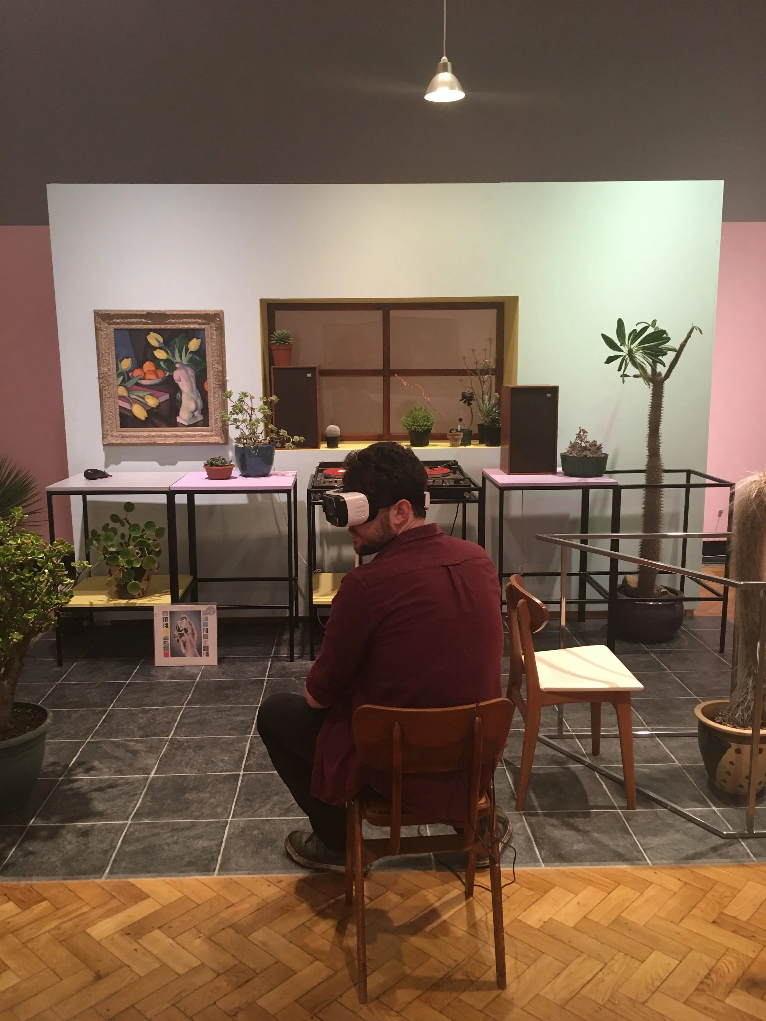  Image description: A man sits in a wooden chair with his back to the camera, wearing a VR headset. In front of him in the physical gallery is a freestanding wall with a window and a painting of a still-life, a few tables with houseplants on, and som