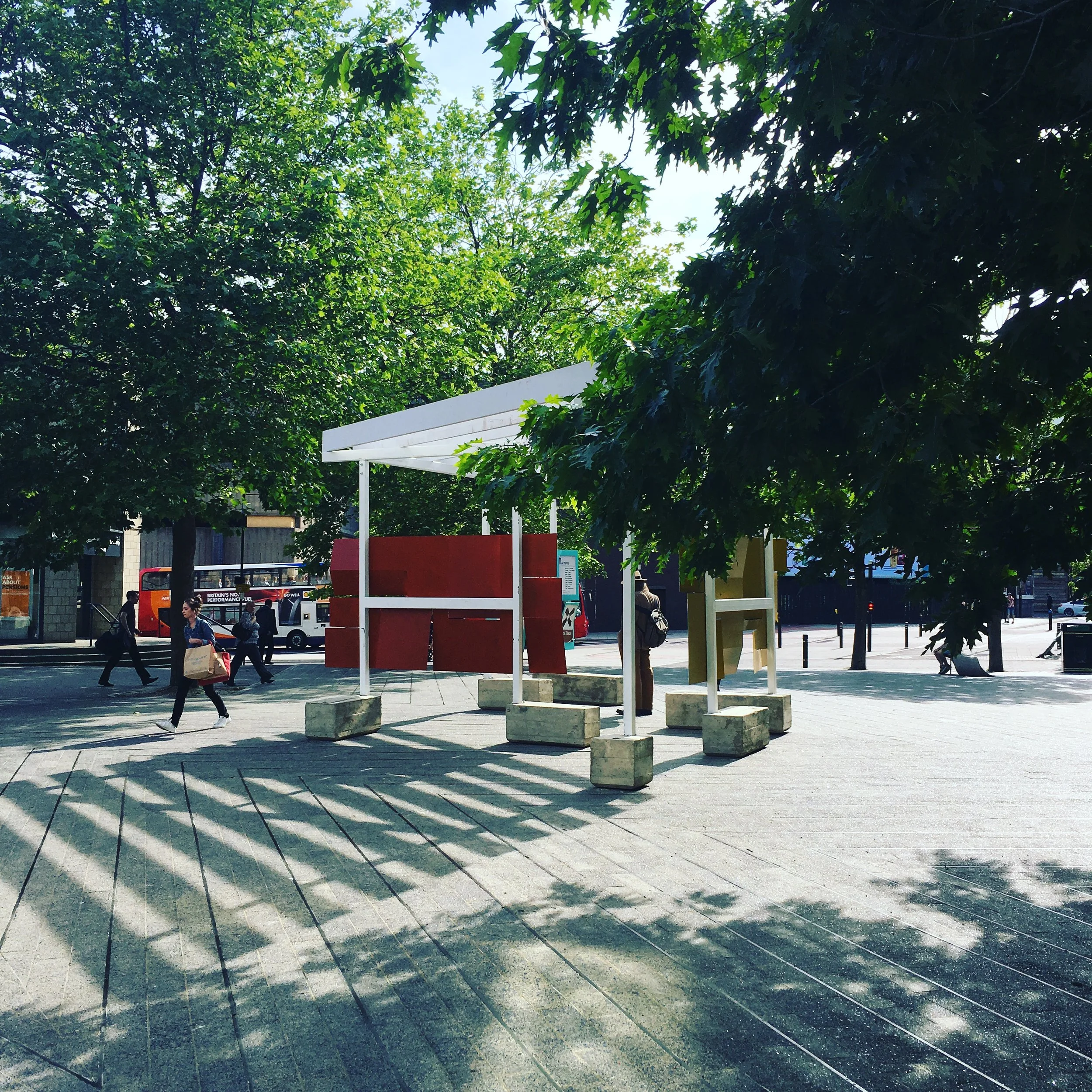  Image description: In a public square in a city centre, with tall trees in the foreground and people and buses passing by in the background, sits a pavilion made of white wood anchored in concrete blocks which double as seats. Mostly open to the ele