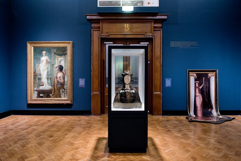  Image description: An install photo of the second room of ‘The Enchanted Interior’ exhibition, with a glass case in the foreground containing a life-size ivory and bronze bust of an eerily life-like ‘Lamia’, by George Frampton. On the wall to the le