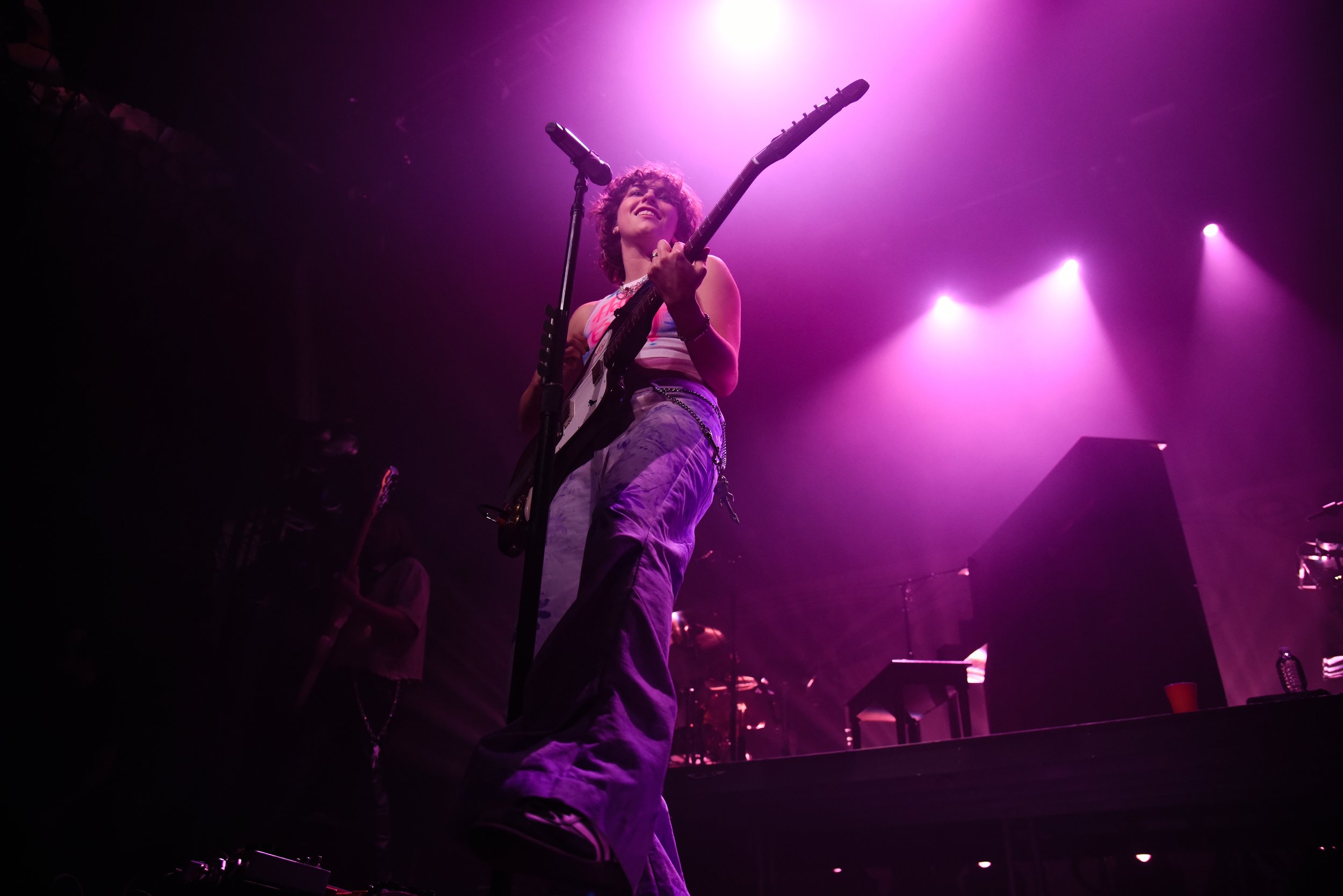  King Princess leans back, kicking out her leg while strumming her guitar. 