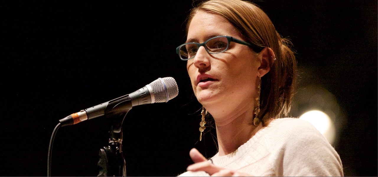 Image: A head shot of Liz speaking into a microphone, wearing a white sweater and long gold dangly earrings.