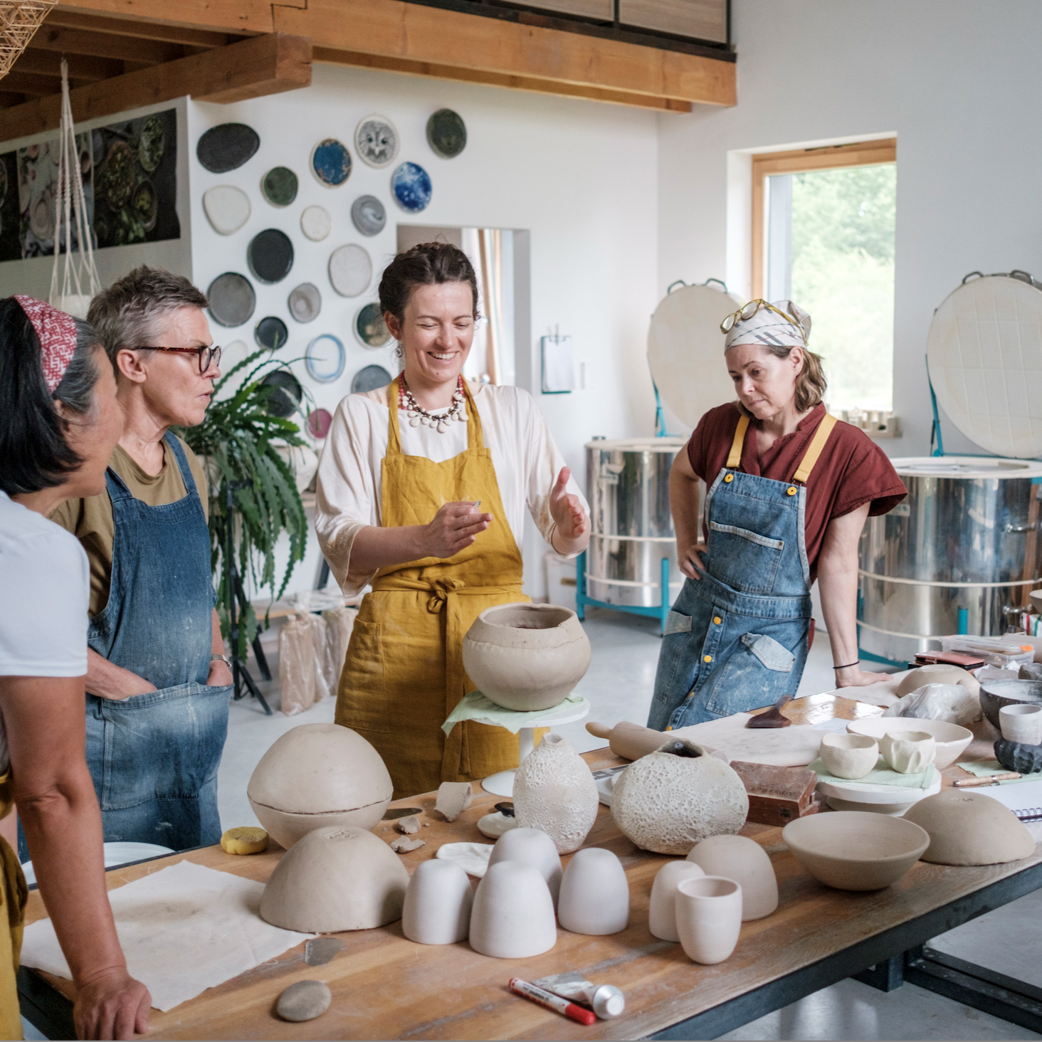 A dining room with a long wooden table set with black and white ceramics and gold utensils, against a white wall with a wooden sideboard and decor, with a blurred person walking in the background.