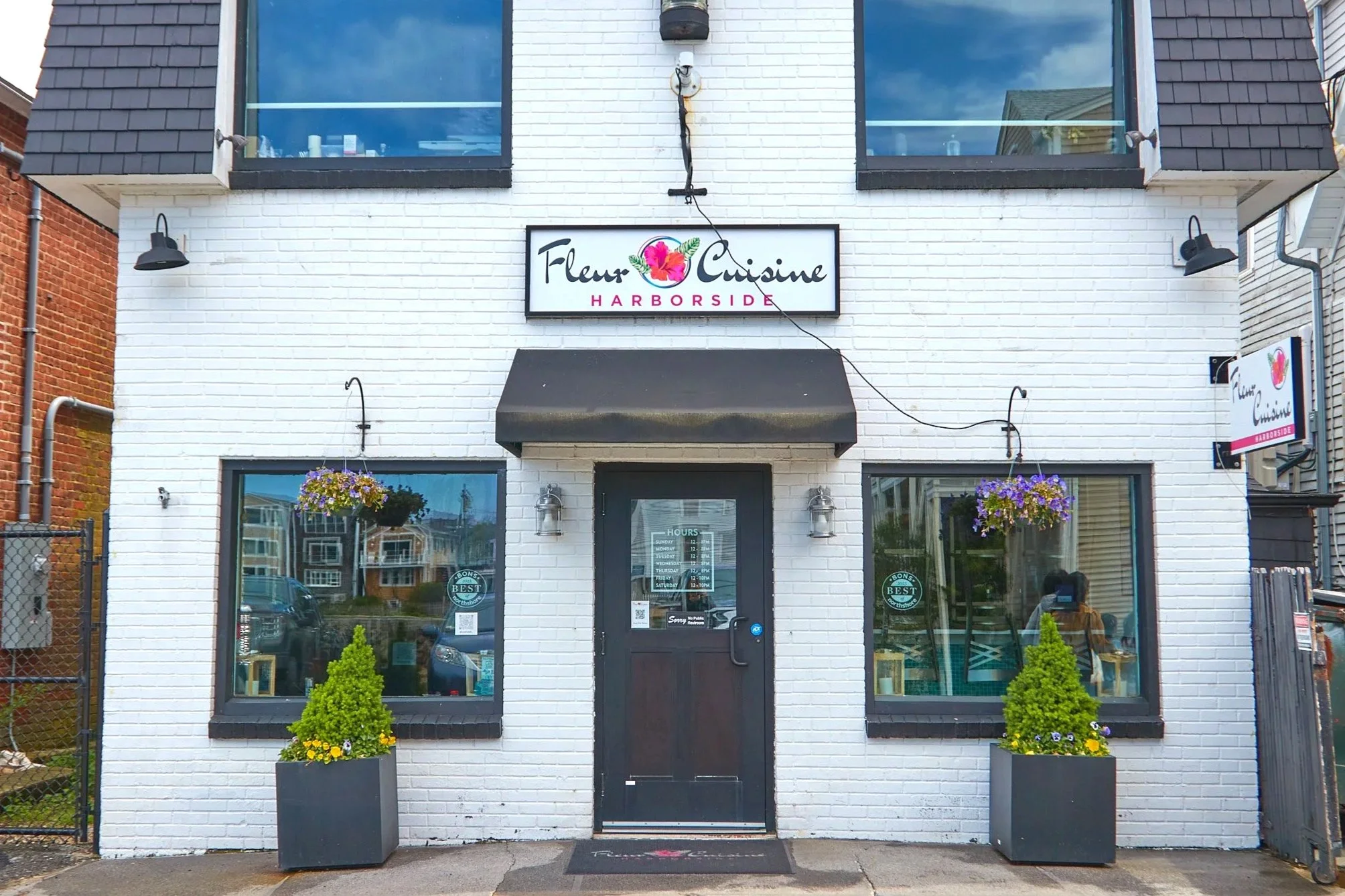 Exterior of Fleur Cuisine Harborside restaurant in Rockport, MA with black awning and hanging flower baskets