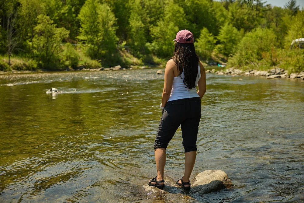 Woman wearing a white shirt, black pants, and red cap standing on a rock overlooking river in Vermont