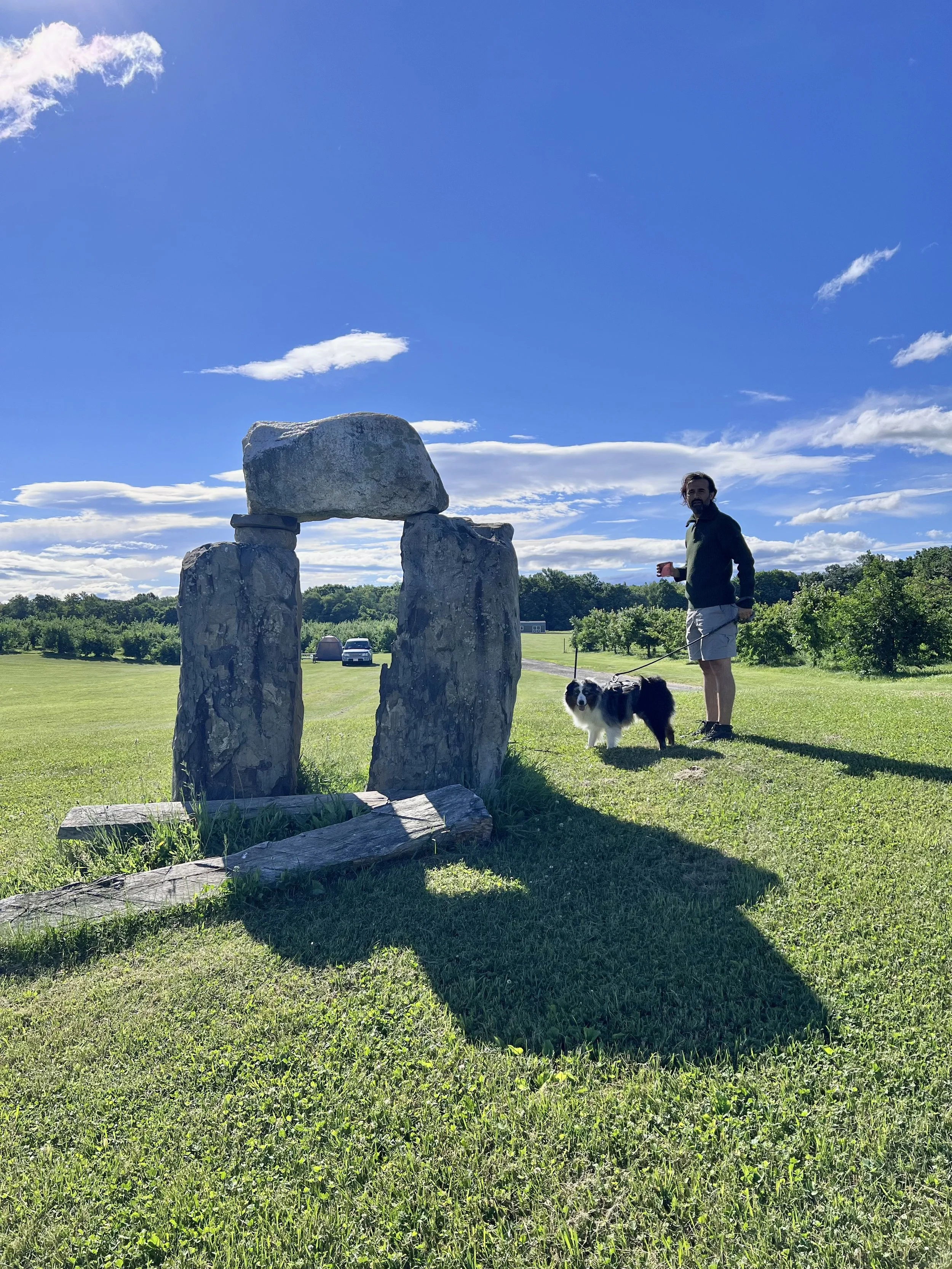 Latino man and dog standing near a rock formation on grass and blue sky in background