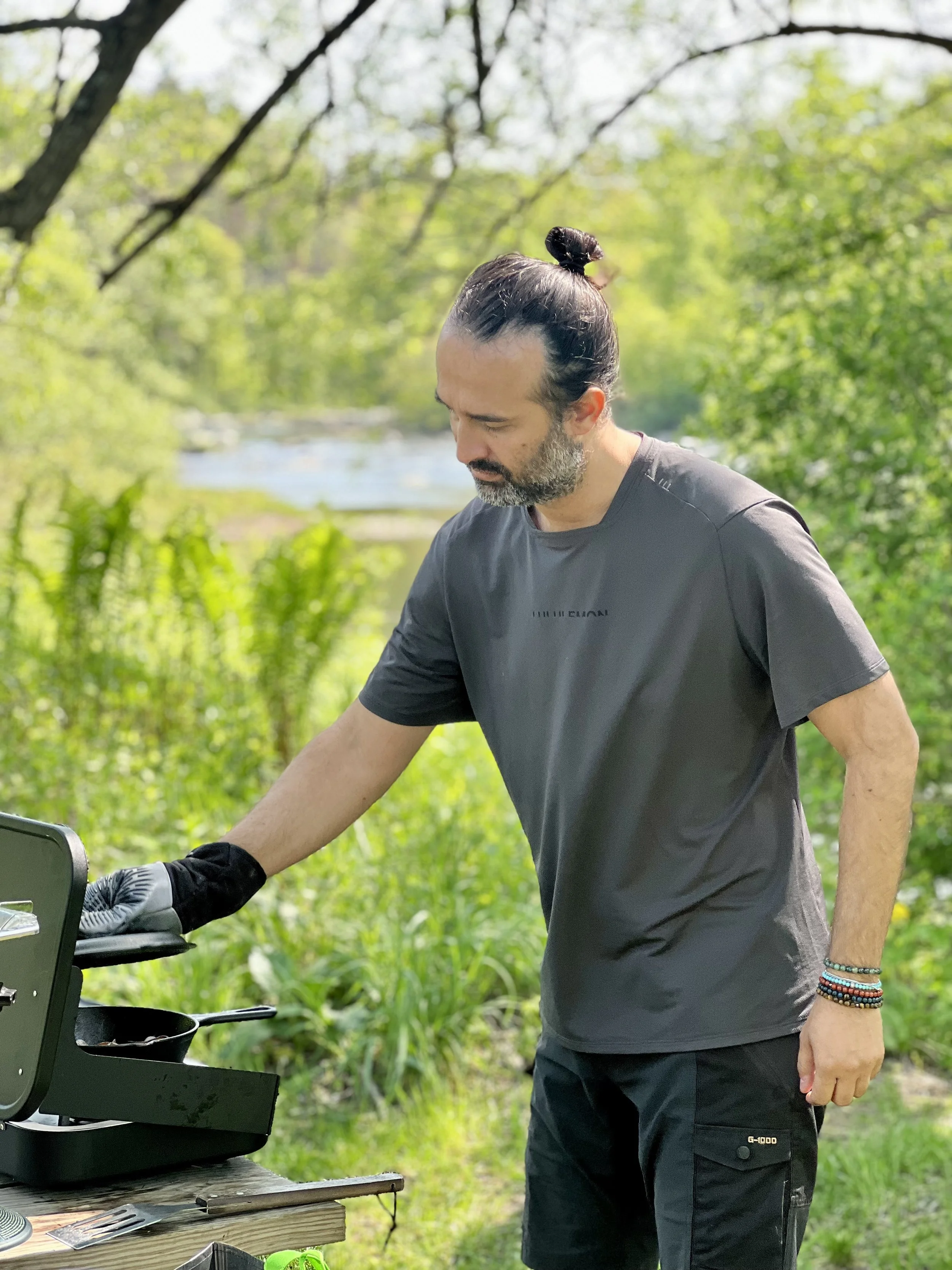 Latino man wearing hair in a bun cooking on a gas camping stove in nature