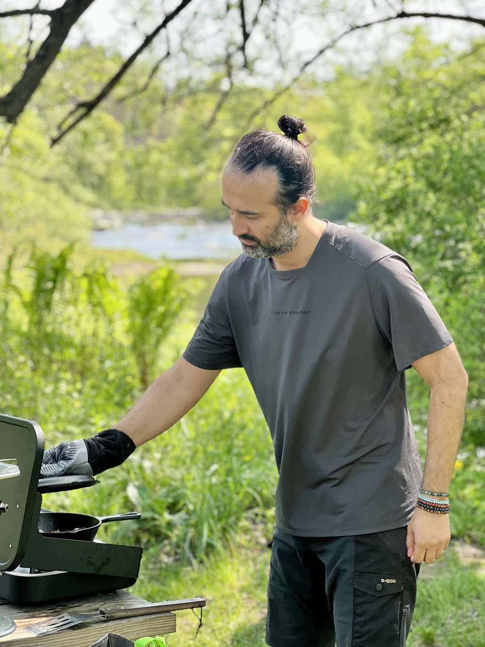Latino man wearing hair in a bun cooking on a gas camping stove in nature