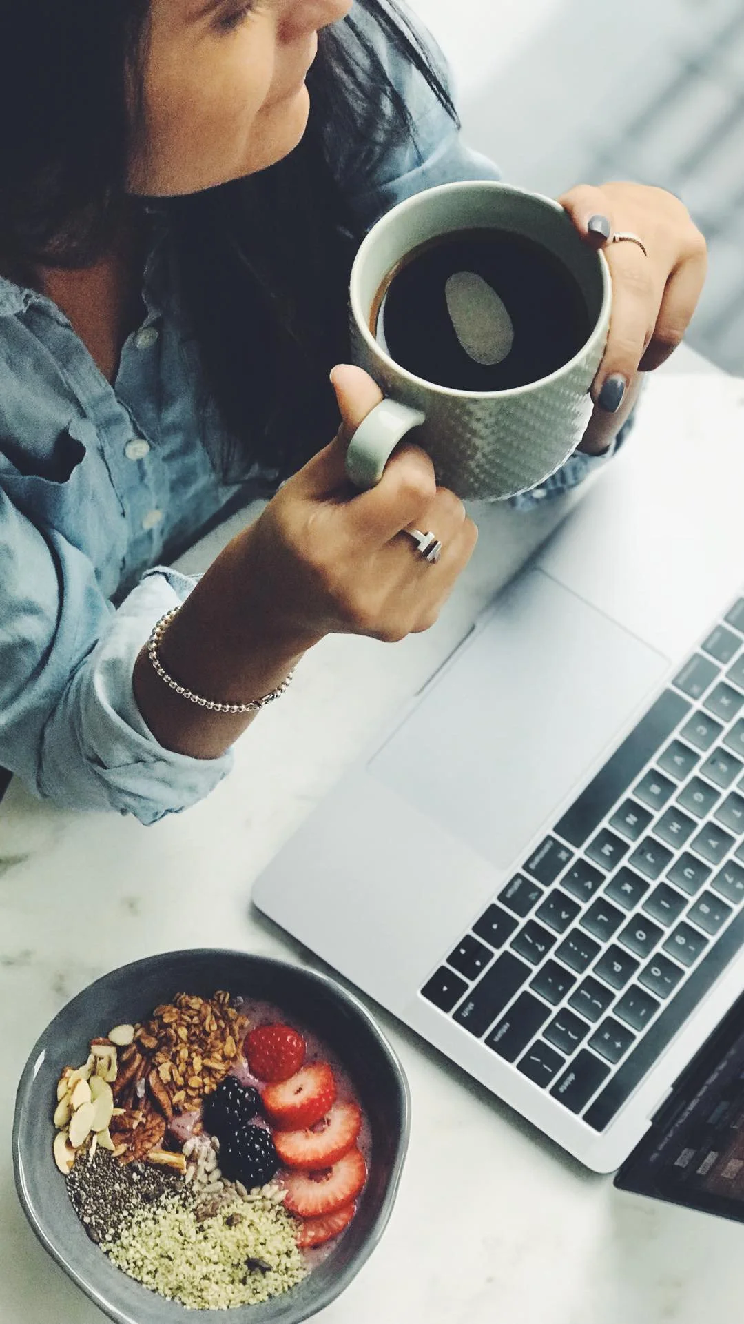 Overhead view of hands holding a coffee mug over a laptop beside a smoothie bowl with fresh berries and granola on a marble surface