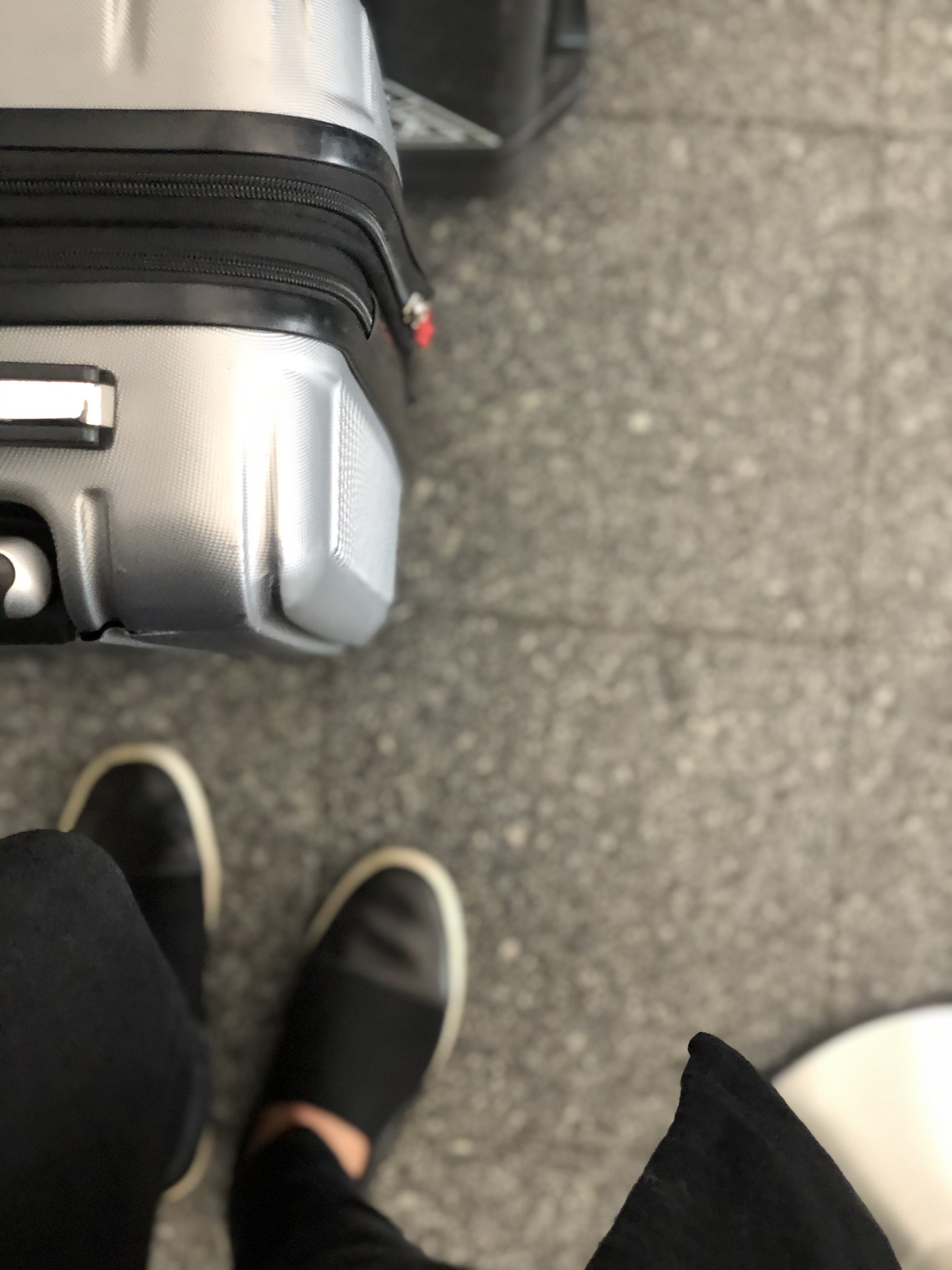 Close-up of a silver hardshell suitcase and sneakers on a grey airport floor