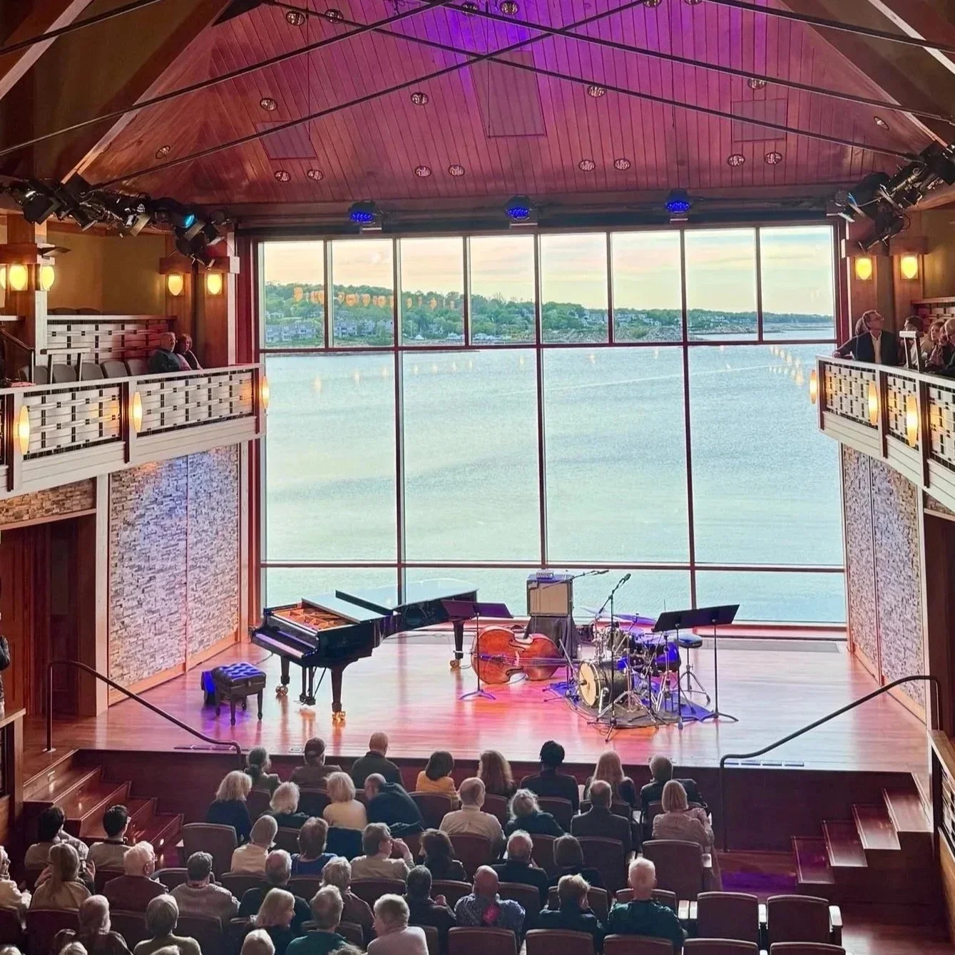 Interior of Shalin Liu Performance Center in Rockport, MA with grand piano and jazz setup on stage, full audience, and harbor visible through floor-to-ceiling window