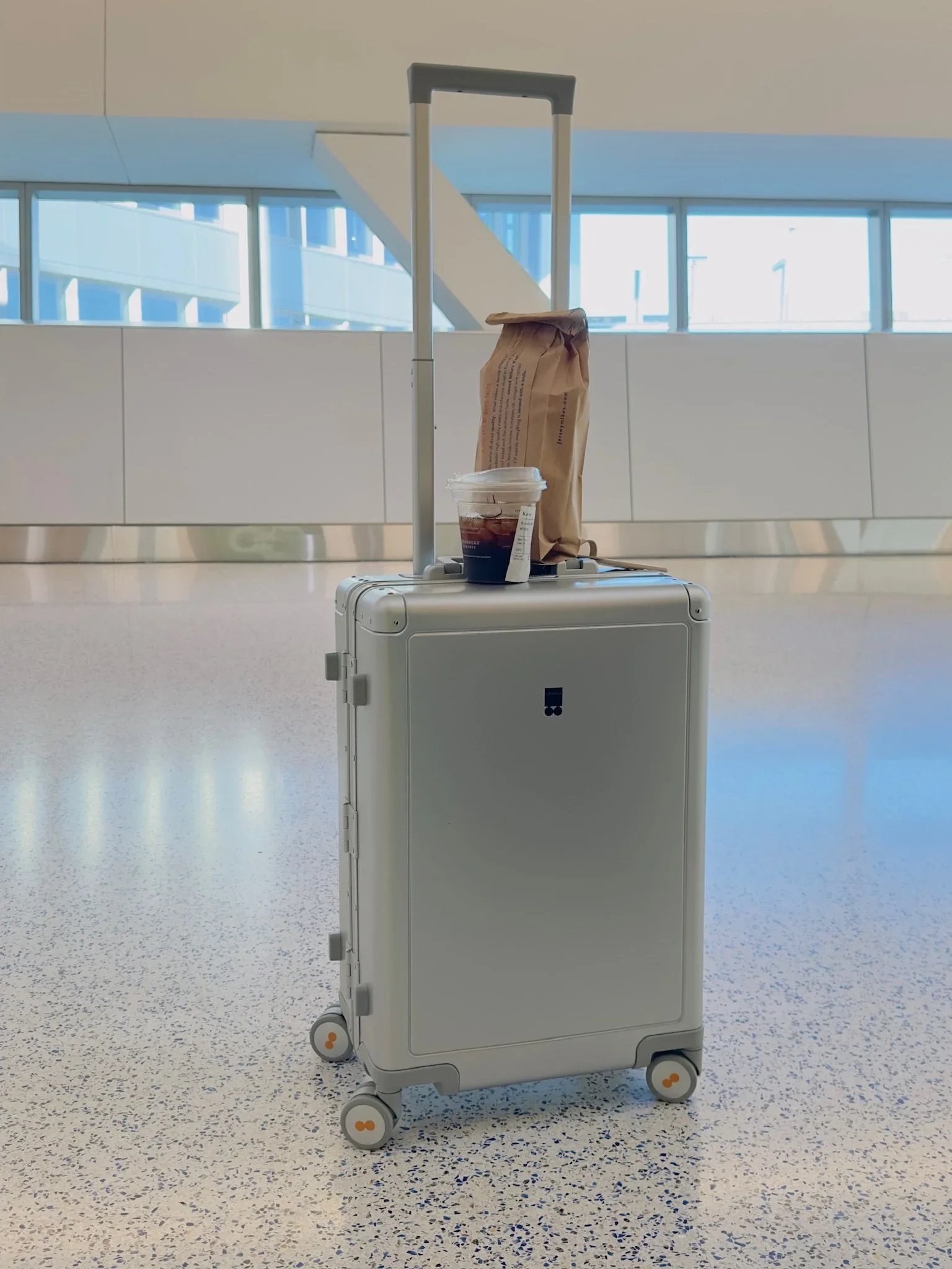 Silver Level8 hardshell suitcase with an iced coffee and paper bag on top in a bright modern airport terminal