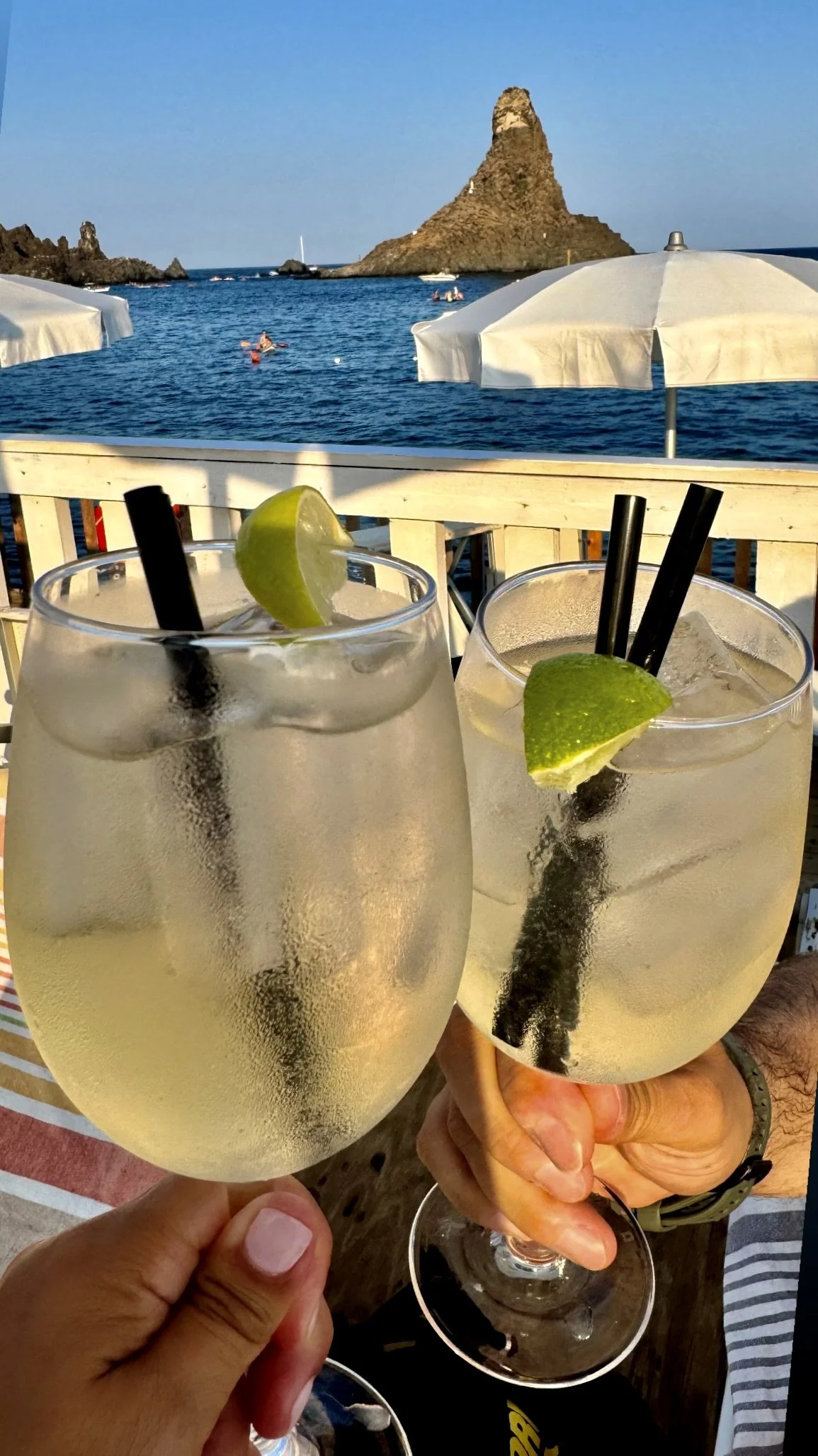 Two iced cocktails with lime garnishes raised in a toast overlooking the sea stacks of Aci Trezza on the coast of Sicily, Italy