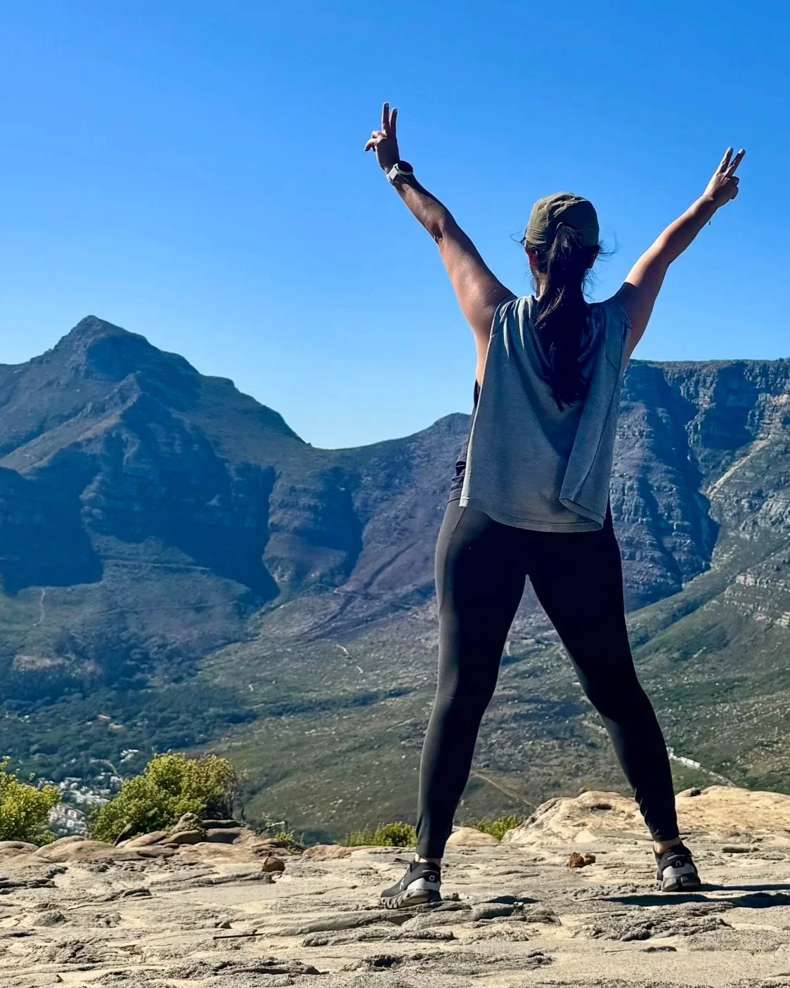 Karen Gomez with arms raised on a rocky overlook above Cape Town with Lion's Head mountain and Table Mountain in the background, South Africa