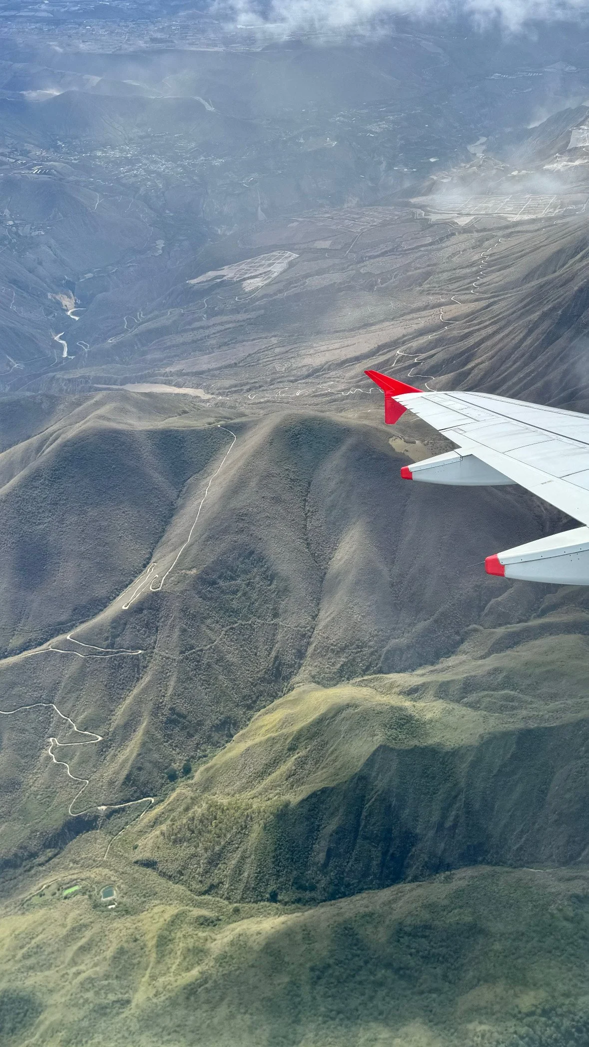 Aerial view of rugged Andes mountain ridges and winding roads from a plane window with a red Avianca aircraft wing visible