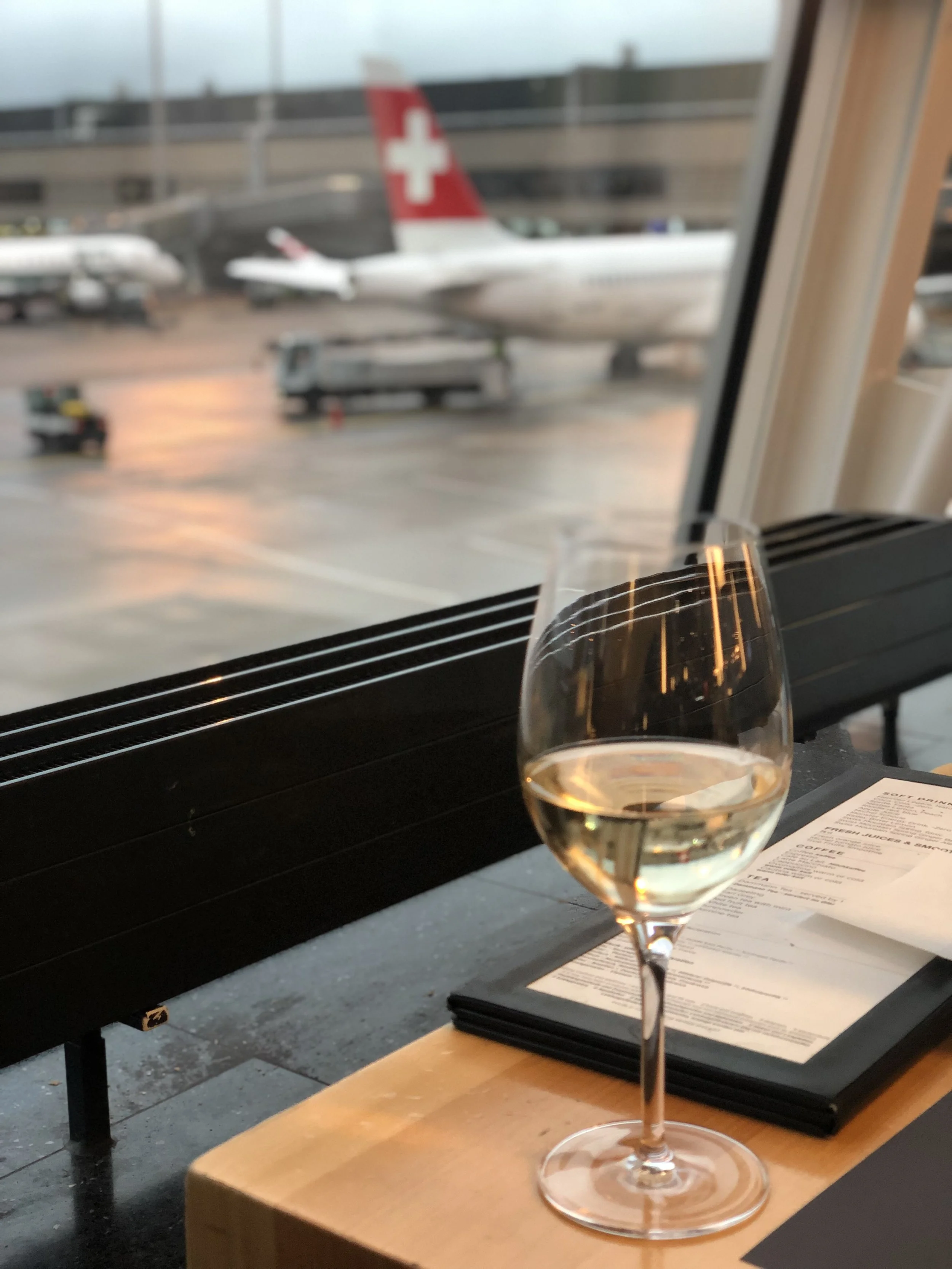 Glass of white wine on a wooden ledge at Zurich Airport with Swiss Air and British Airways planes visible on the tarmac