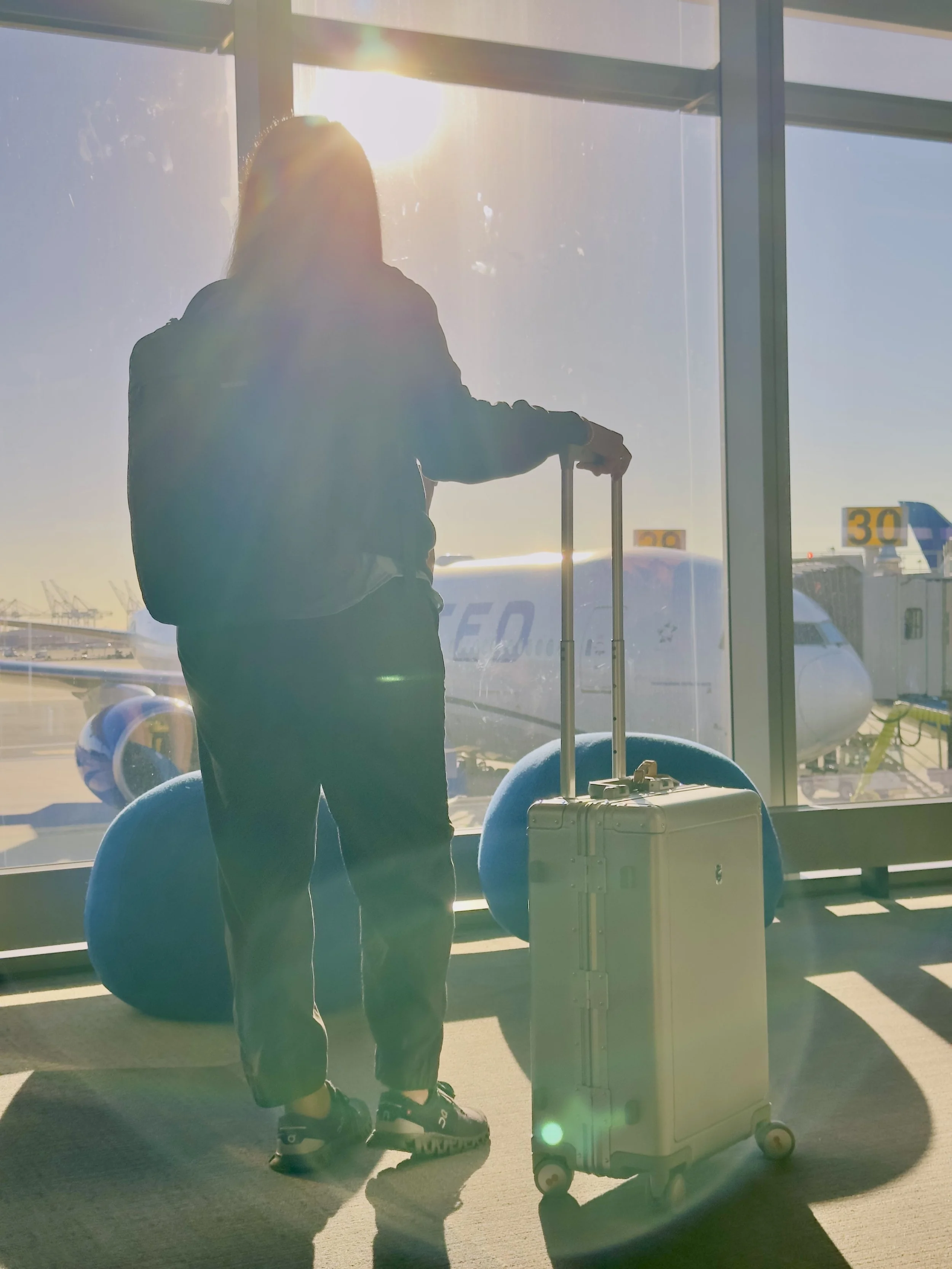 Silhouette of a woman standing with luggage at an airport gate window with a United Airlines plane on the tarmac backlit by sunlight