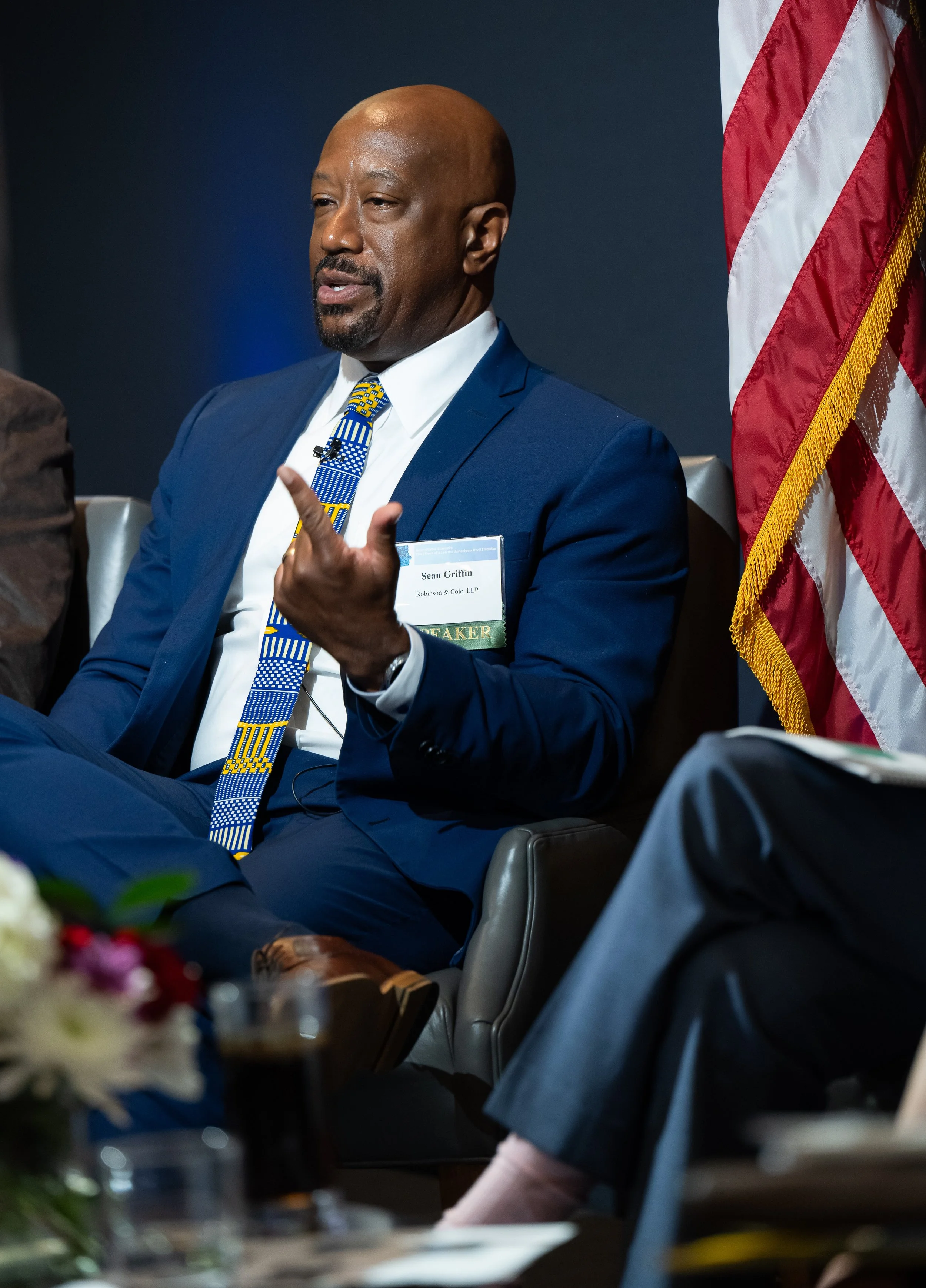 Sean C. Griffin in a blue suit and patterned tie speaking on artificial intelligence and litigation, with an American flag beside him.