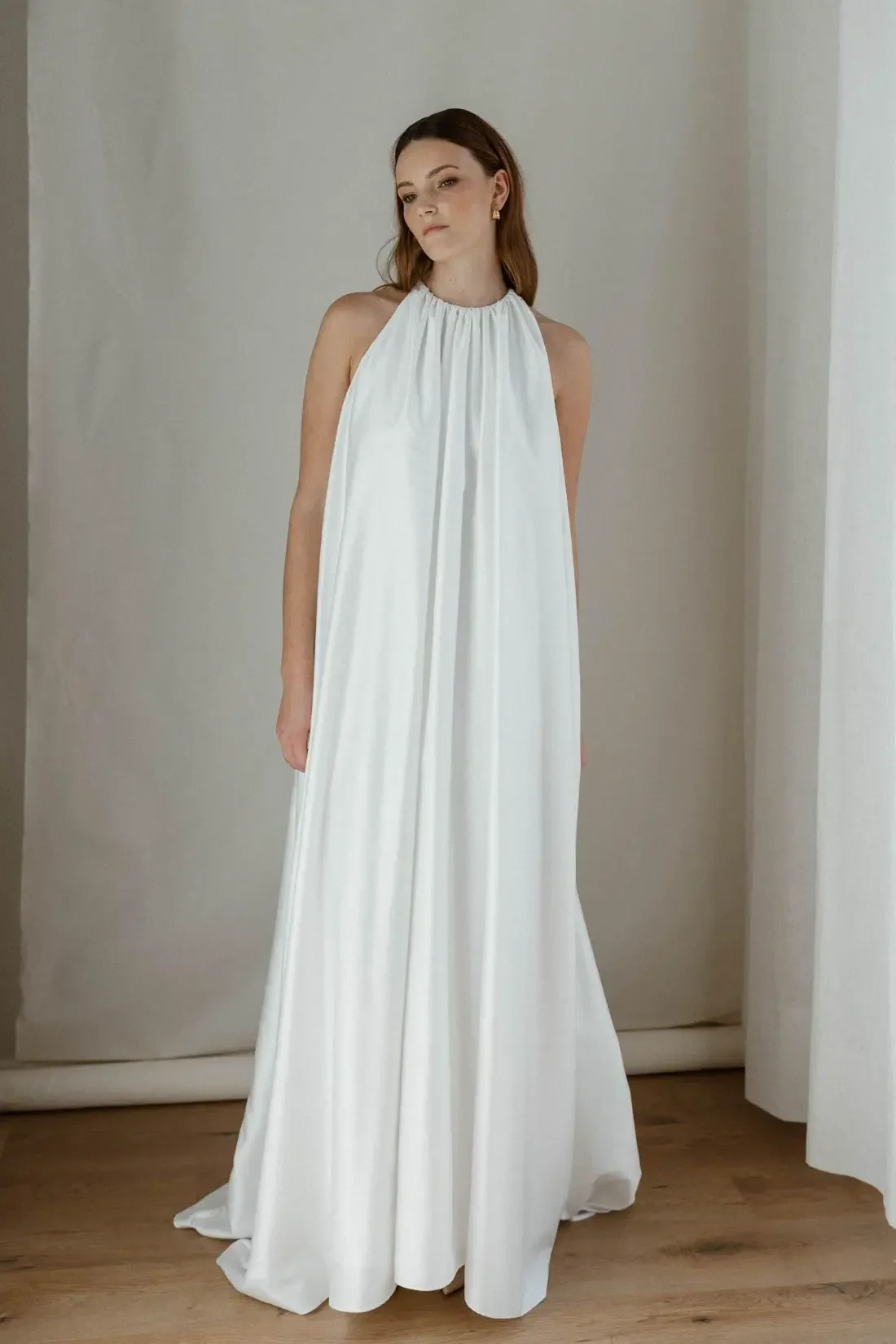 A woman with brown hair in a sleeveless white gown with high neckline, standing in front of a white wall and curtains, with light wood flooring.