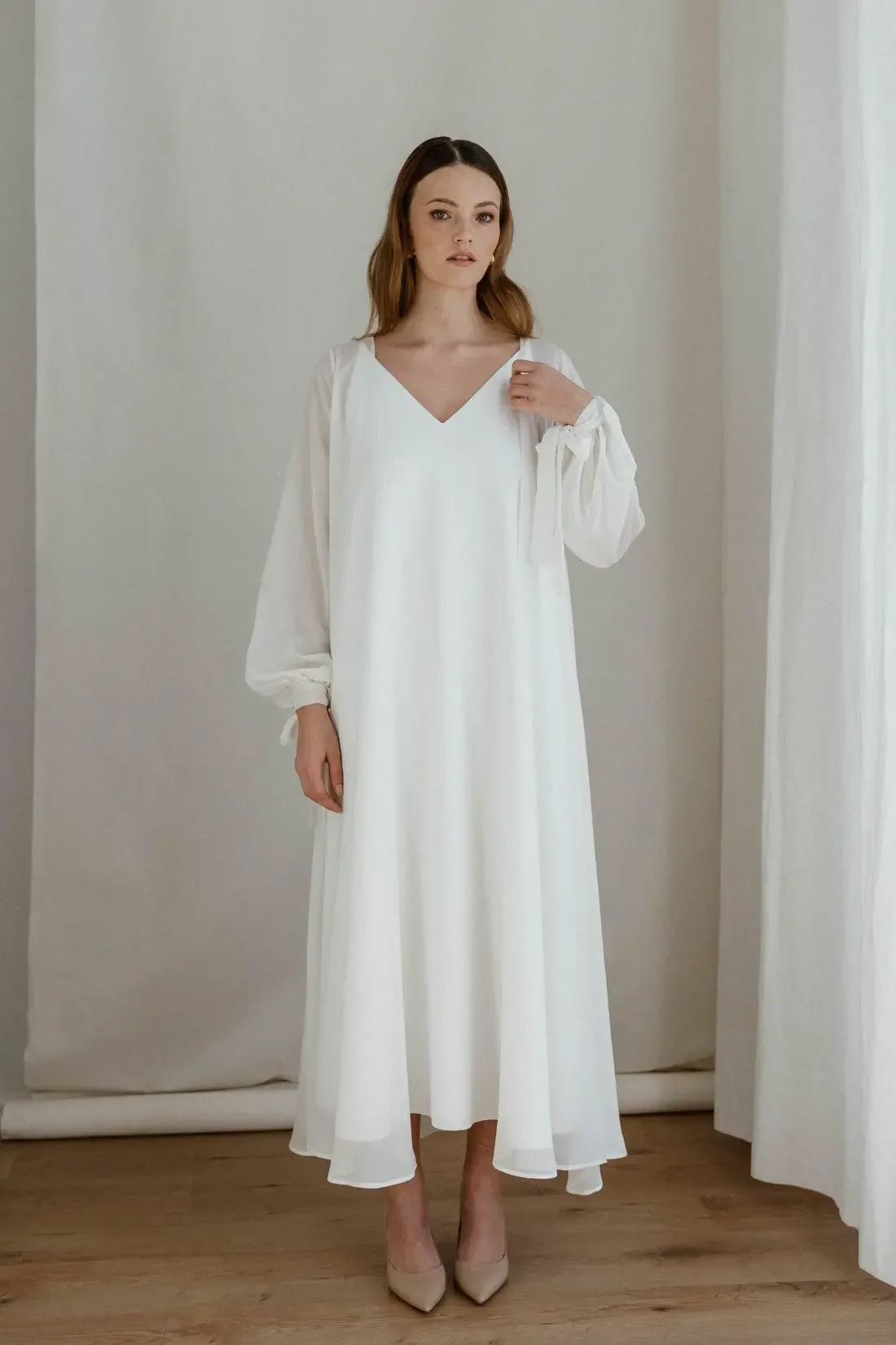 A young woman is standing in a room, wearing a long white dress with long, billowy sleeves and beige high heels, near cream-colored curtains and a plain wall.