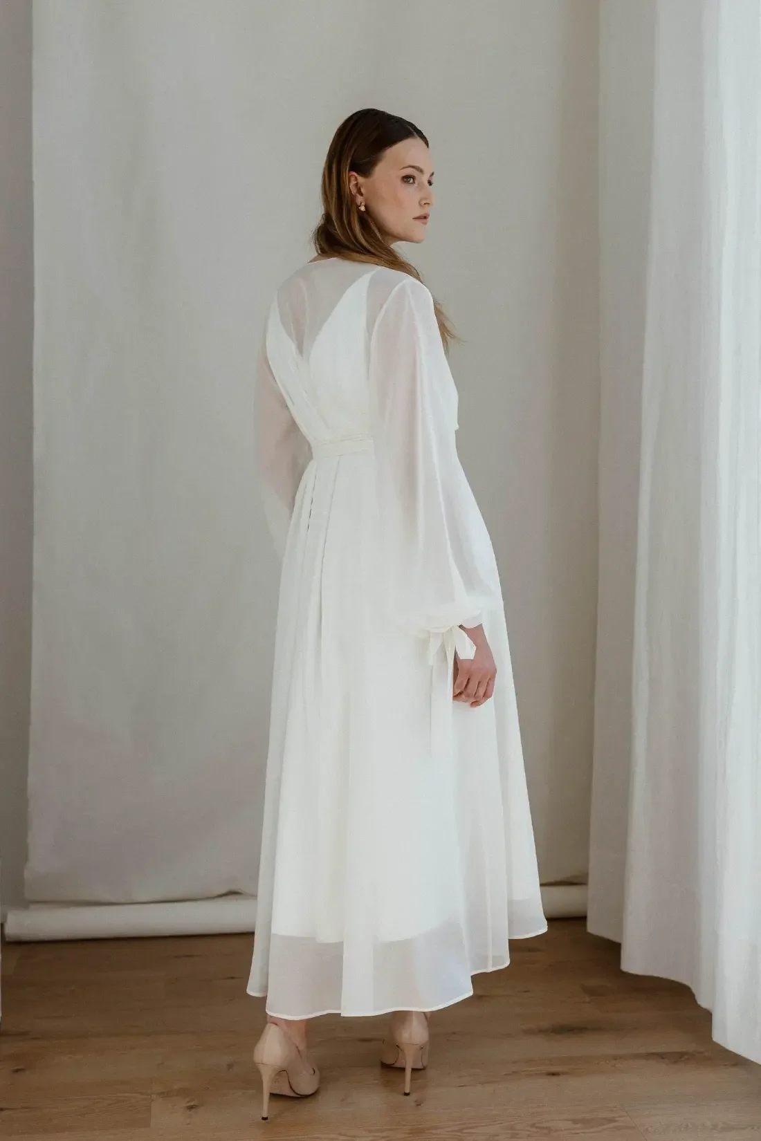 A woman in a white dress standing indoors near white curtains.