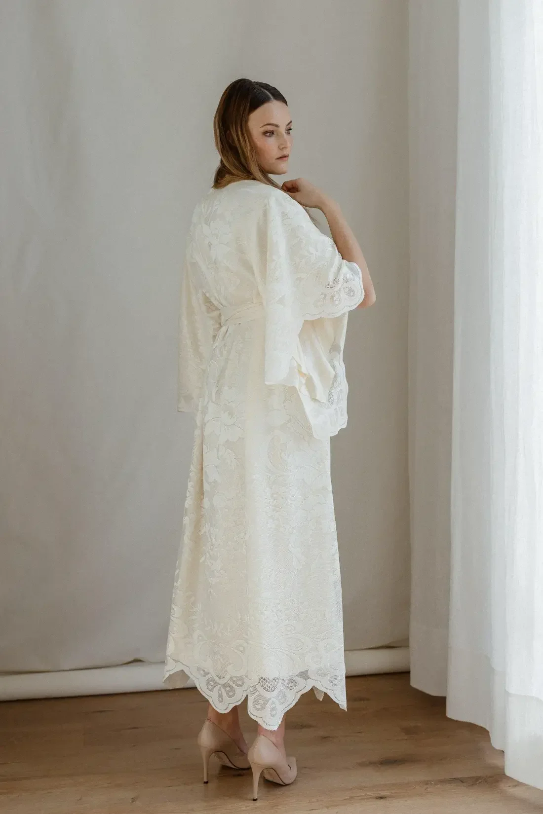 A woman with brown hair stands in a softly lit room, wearing a white lace dress with long, flowing sleeves, and beige high heels, as she looks to her left near a curtain.