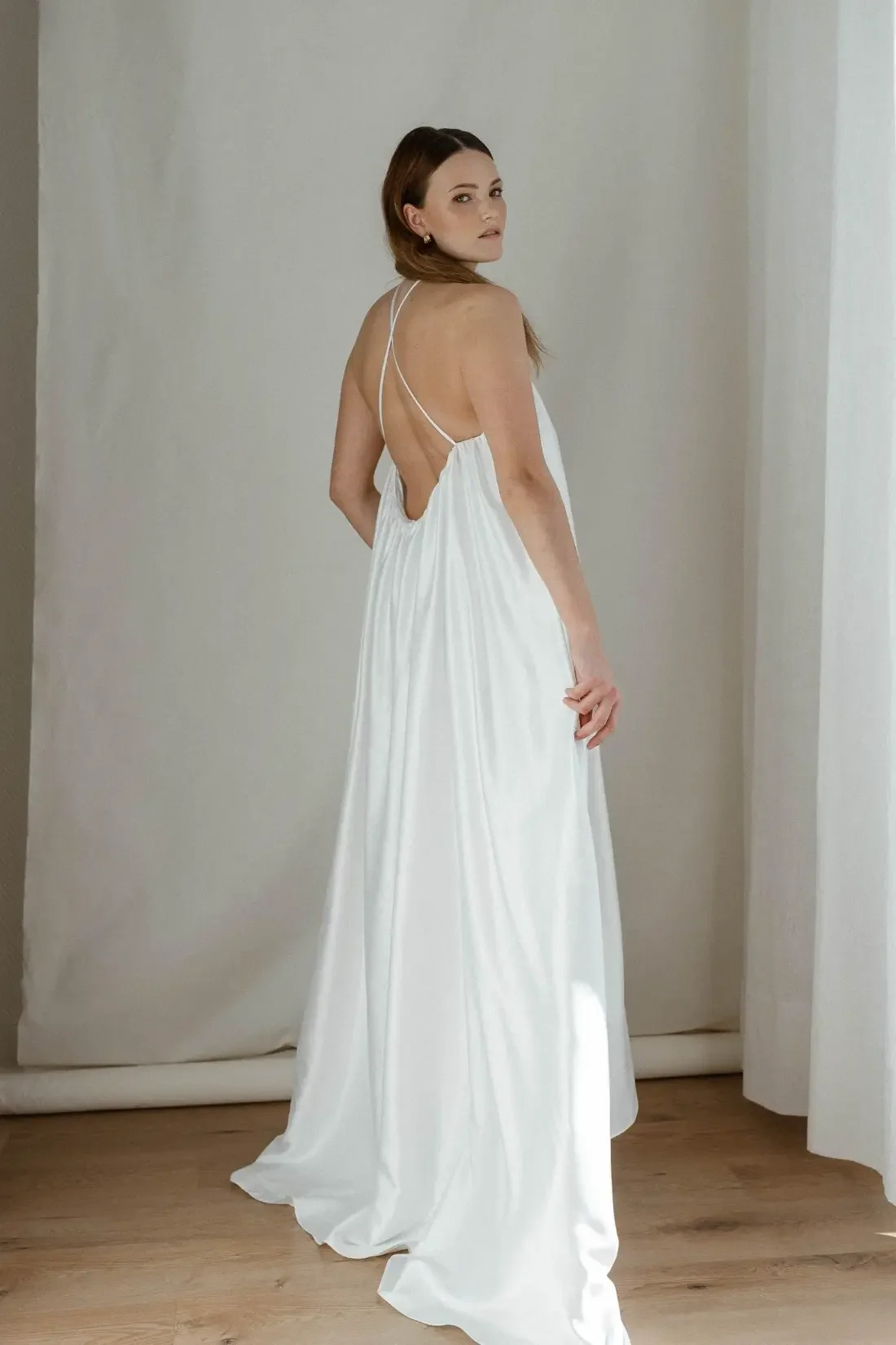 A woman with brown hair in a white backless dress standing on a wooden floor against a plain wall.