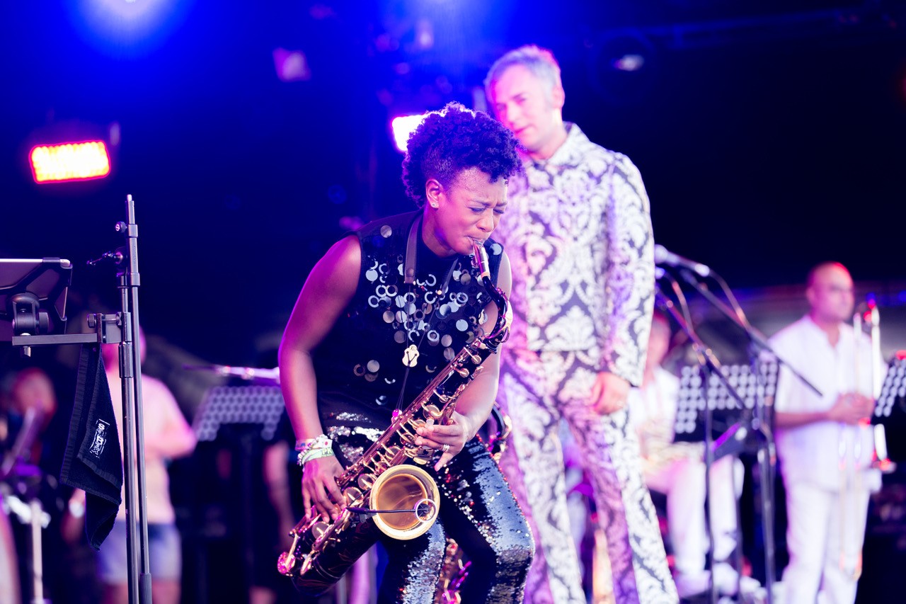  A lady plays saxophone on stage, a man looks on behind 