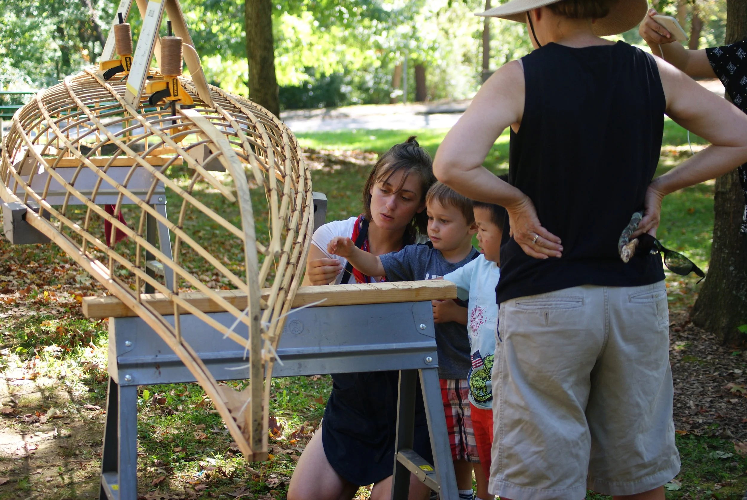 A woman and three children examining a model of a boat or ship under construction outdoors in a park or wooded area.