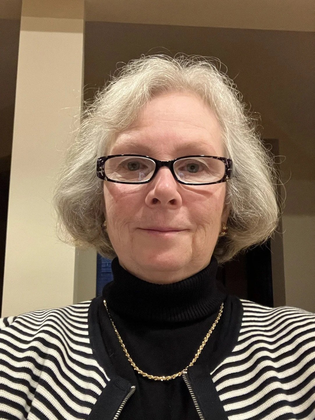 A woman with short gray hair, wearing glasses, a black and white striped sweater, a black turtleneck, a gold chain necklace, and small hoop earrings, taking a selfie indoors.