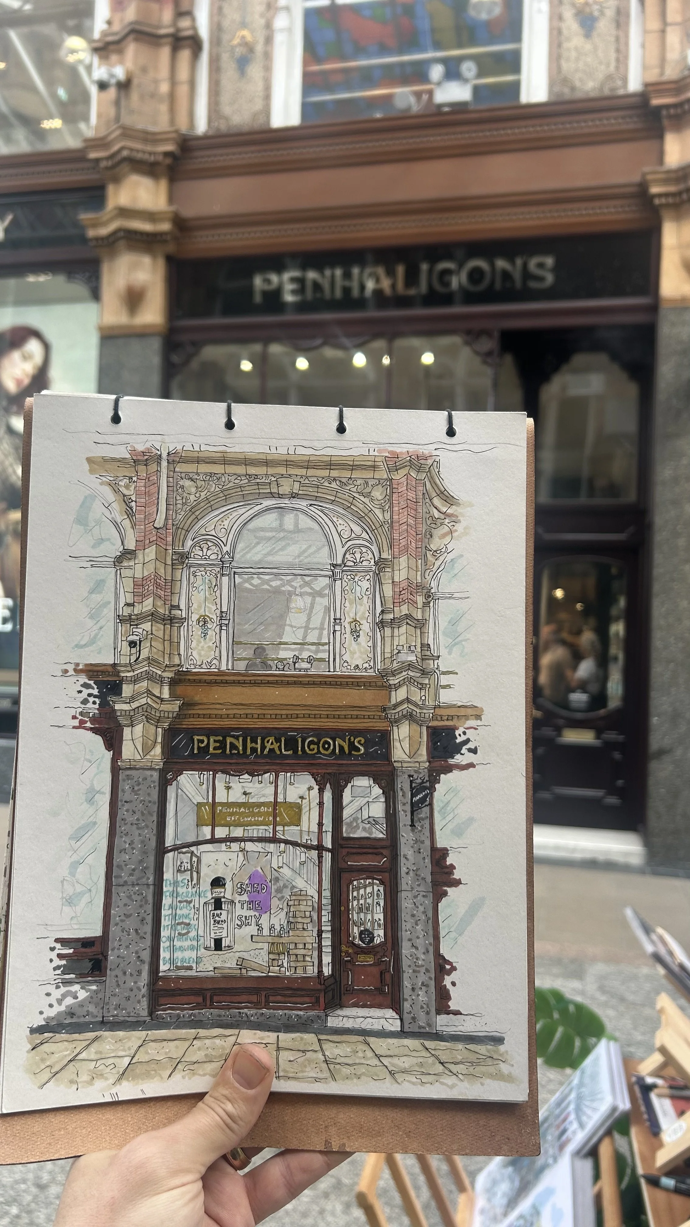 an artist's drawing of the storefront of Penhaligon's, with detailed architectural elements and signs, held in front of the actual Penhaligon's store on the street.