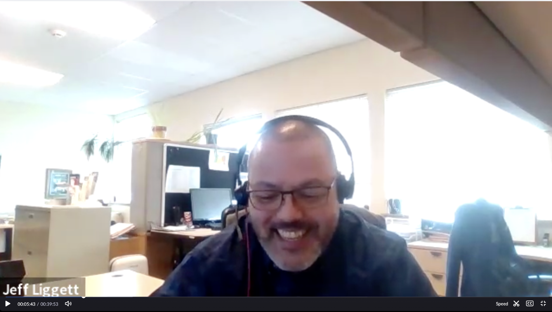 Man with headphones and glasses sitting at desk on Zoom call with office in background